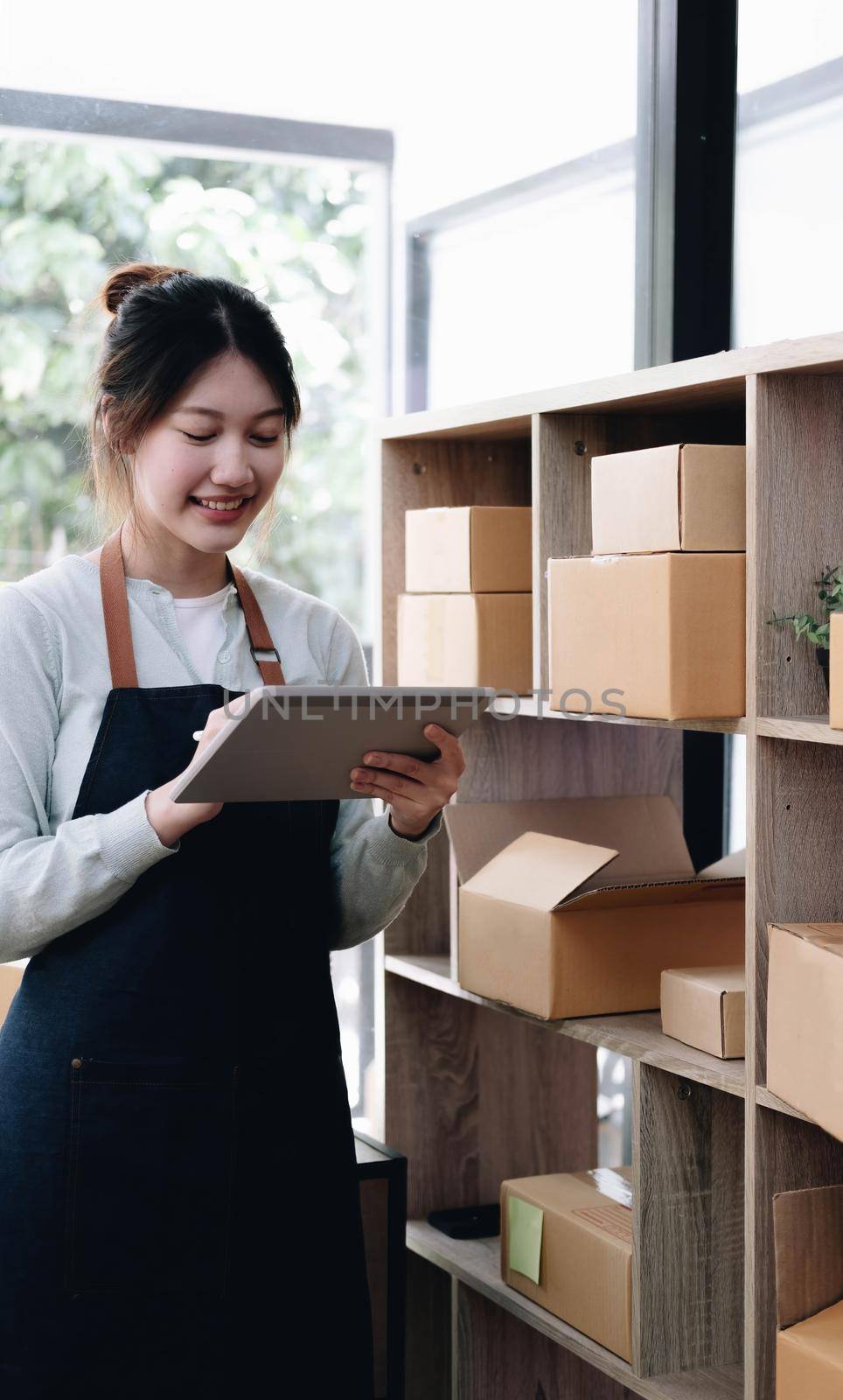 Young beautiful happy asian business woman owner of SME online using laptop receive order from customer with parcel box packaging at her startup home office, online business seller and delivery by wichayada