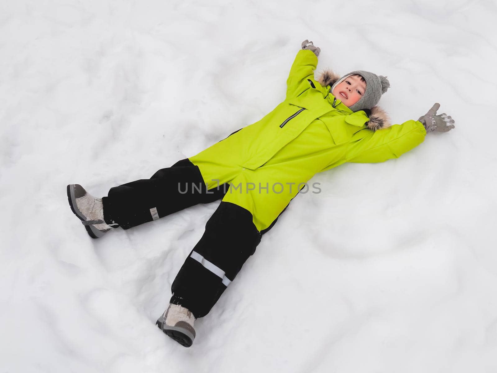 Smiling boy in green jumpsuit is making snow angel shape on snow. Joyful child playing outdoors in snowy weather. Top view on happy kid in colorful overall suit.