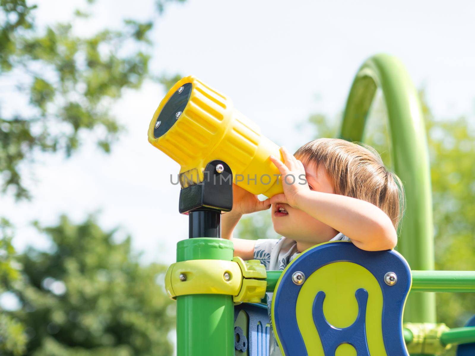 Little kid looks in toy telescope on outdoor playground. Leisure activity for curious children. Summer fun.