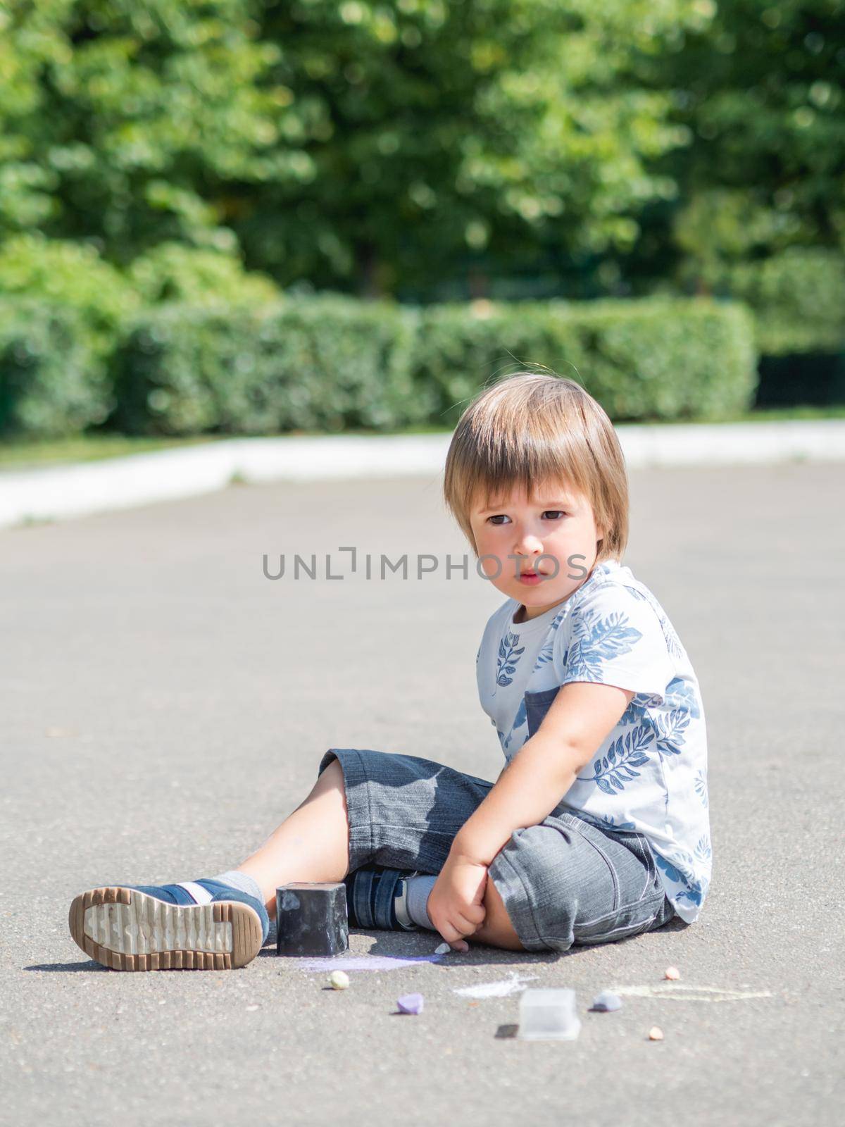 Little mindful kid sits on pavement in park. Kid is drawing something on asphalt. Creative leisure activity for children.