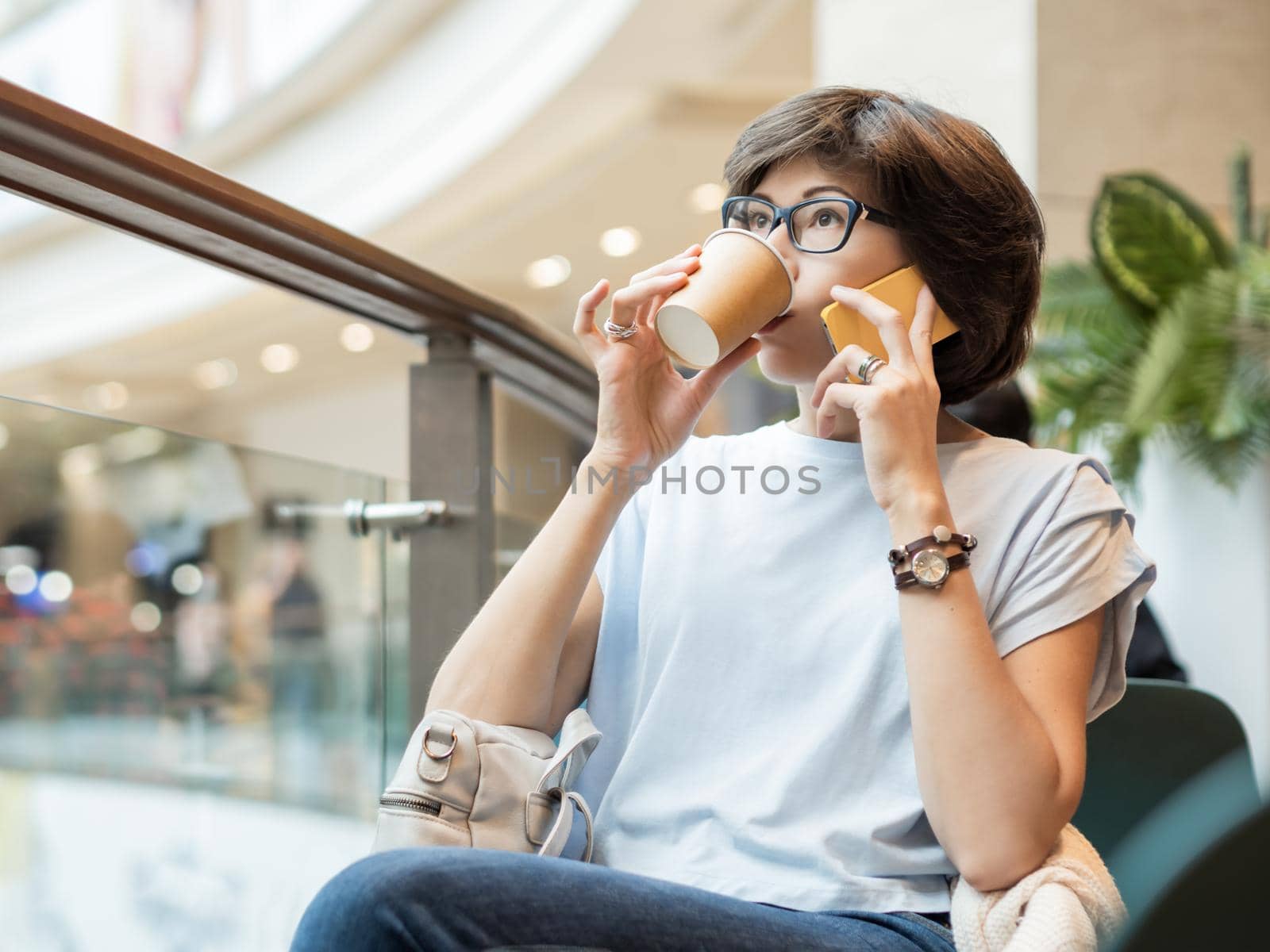 Woman is talking by smartphone and drinking coffee at shopping mall. Coffee break at store. by aksenovko