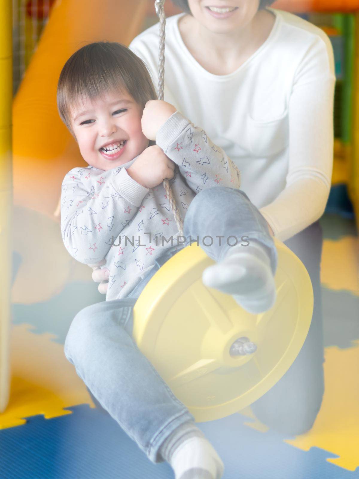 Toddler plays on rope swing with his mother or babysitter. Physical development for little children. Interior of kindergarten or nursery.