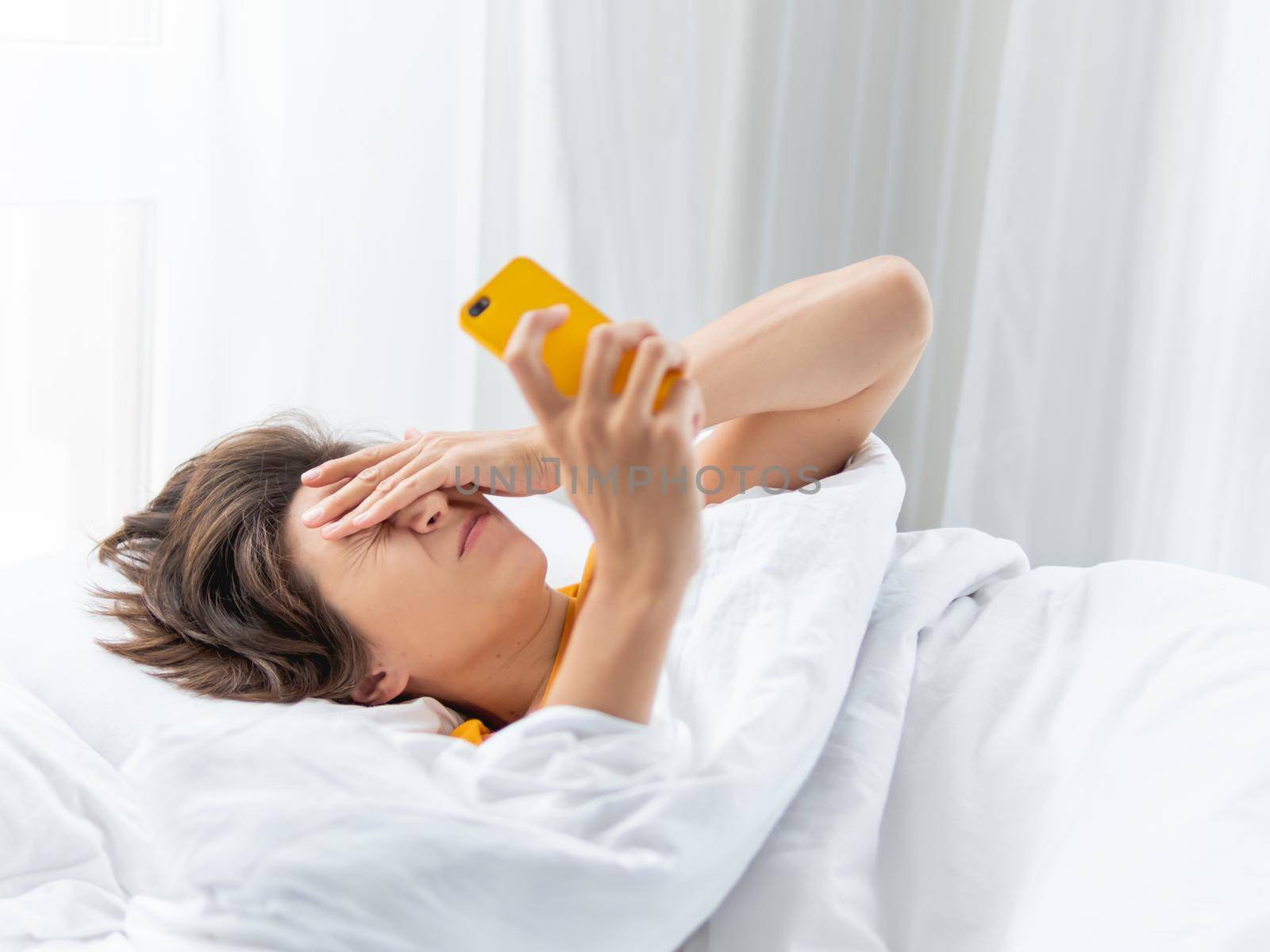 Sleepy woman looks on smartphone screen. Using cellular phone as alarm clock. Wireless technology in bed.