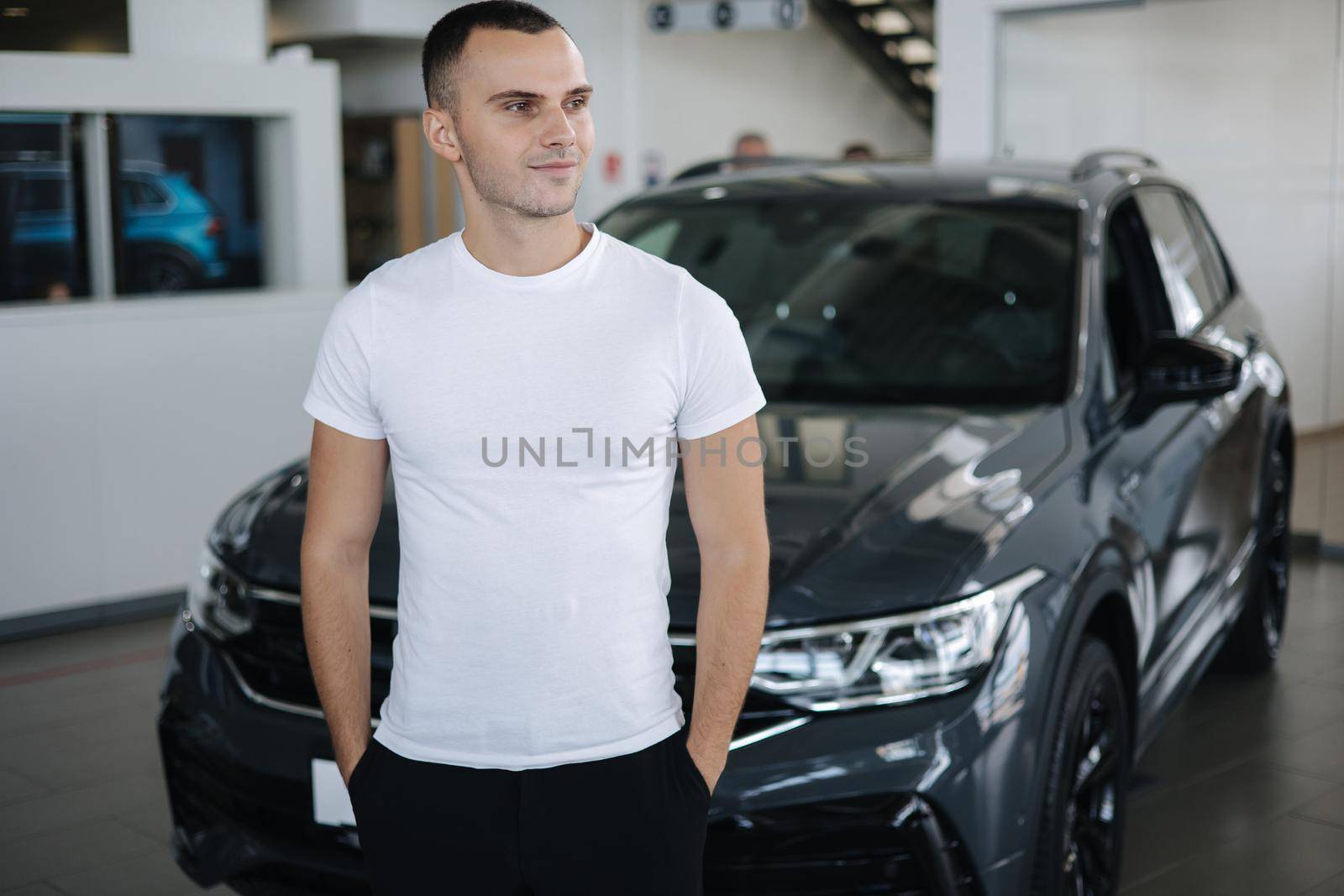 Portrait of handsome man standing by car in car shoowroom. Man buying new car. Male in white t-shirt. Happy person.