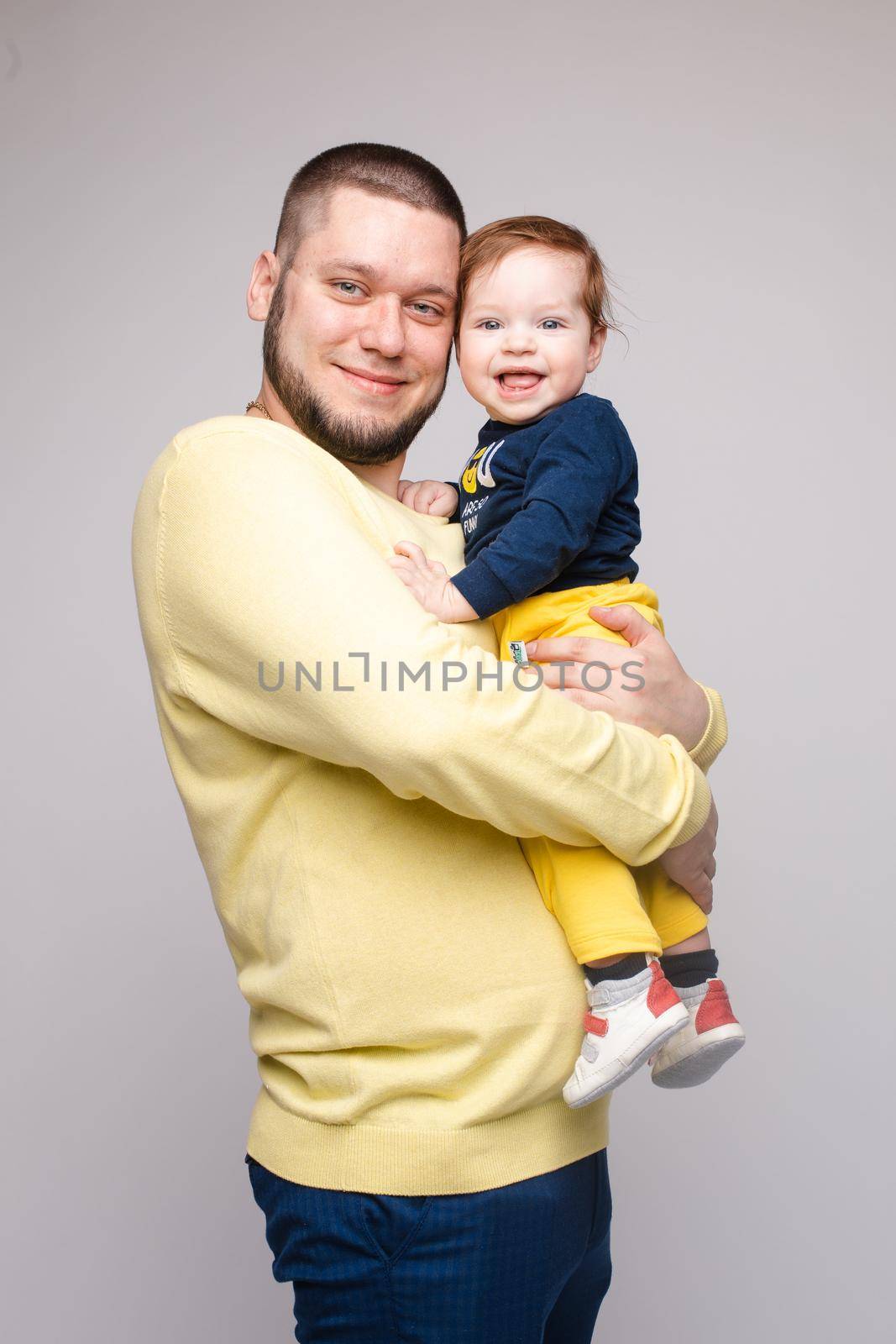 Portrait of happy father in yellow sweater posing with his lovely smiling child. Athletic and young men with beard holding red haired little boy. Handsome brunette dad hugging sweet and cute son.