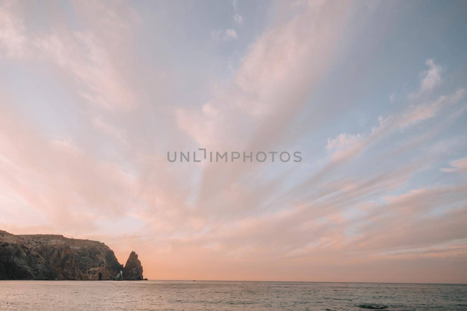 A red burning sunset over the sea with rocky volcanic cliff. Abstract nature summer or spring ocean sea background. Small waves on golden warm water surface with bokeh lights from sun. by panophotograph