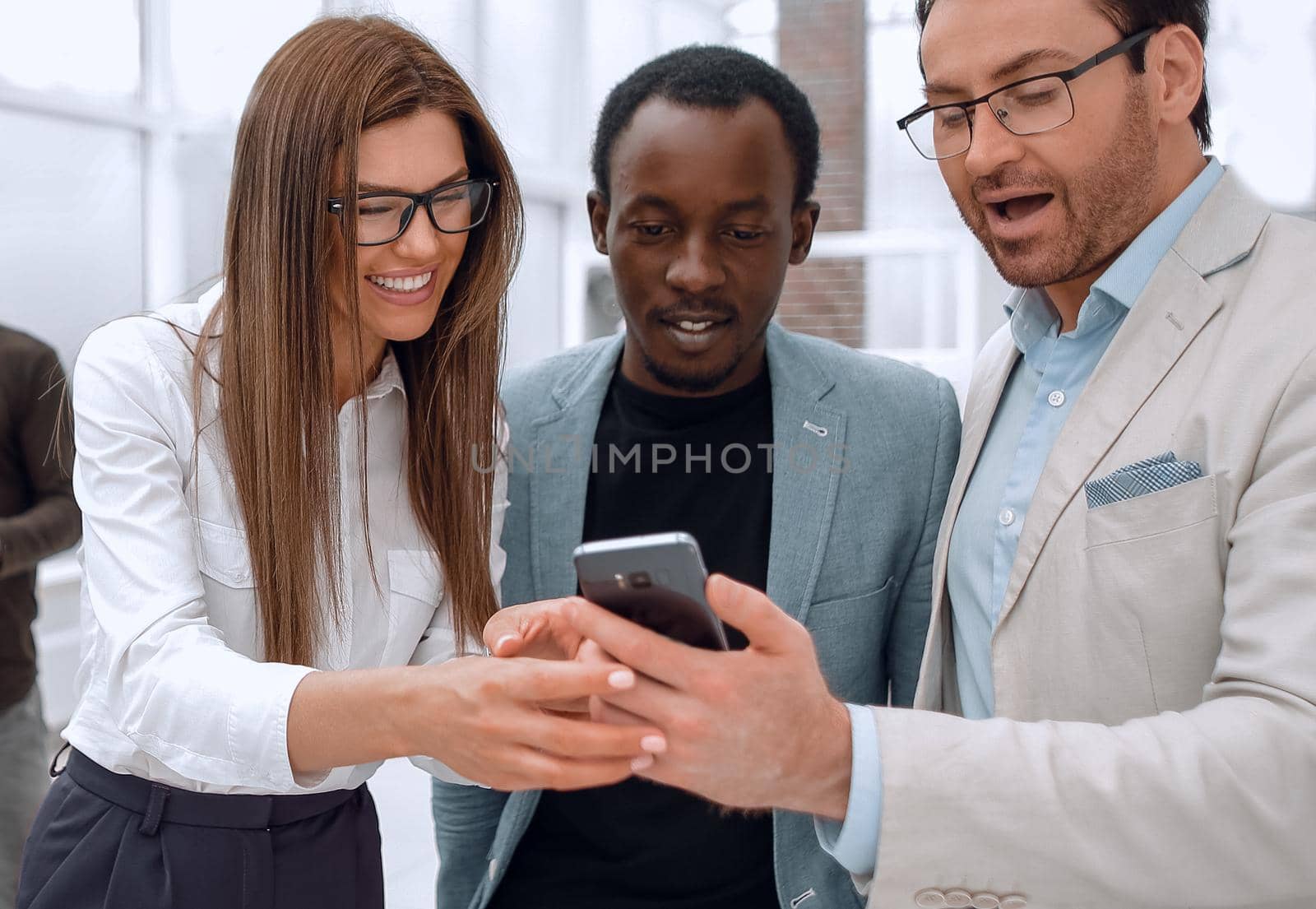 smiling colleagues looking at the smartphone screen, standing in a modern office