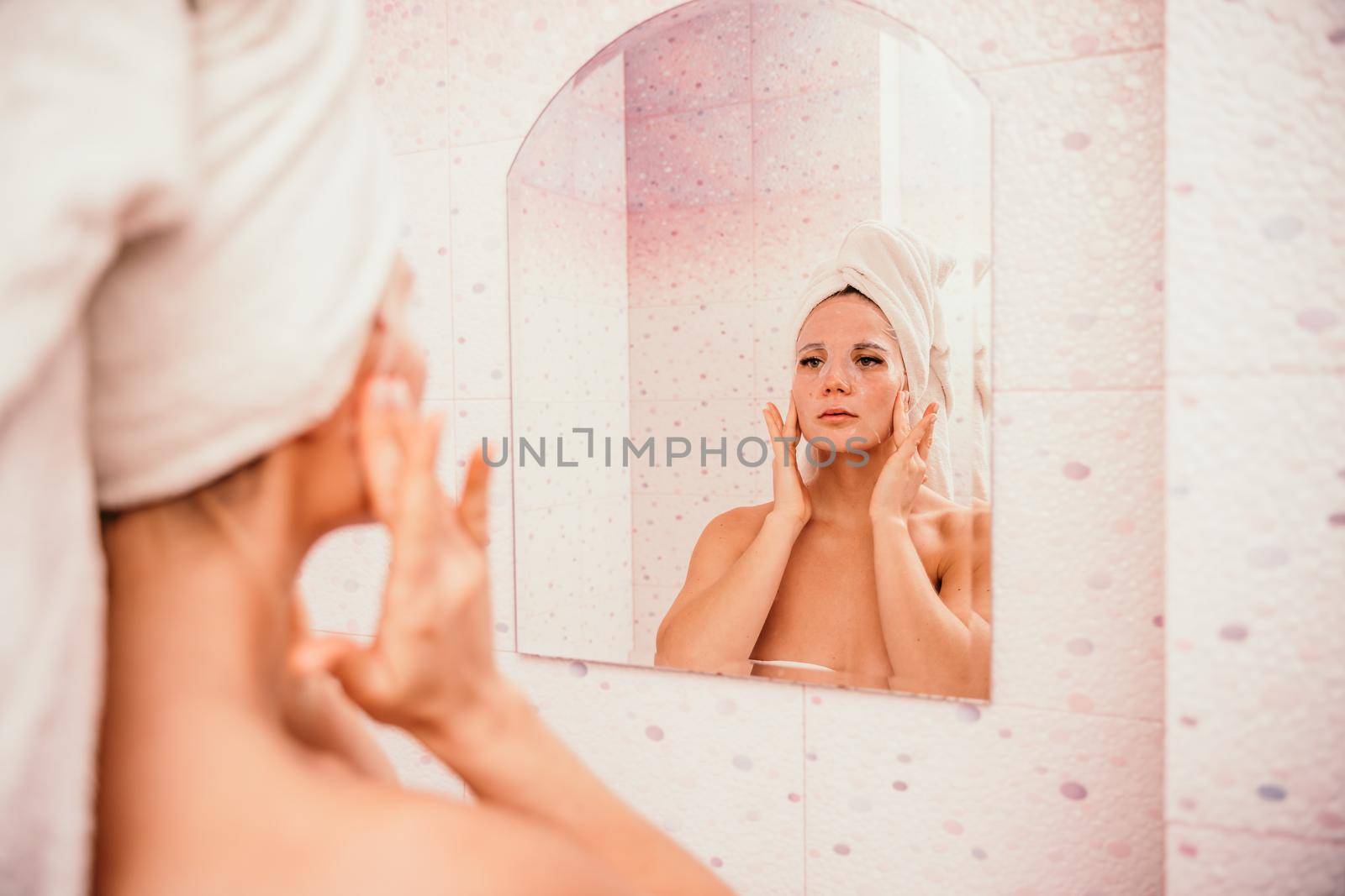 Young beautiful woman using a moisturizing facial mask after taking a bath. Pretty attractive girl in a towel on her head stands in front of a mirror in a home bathroom. Daily hygiene and skin care.