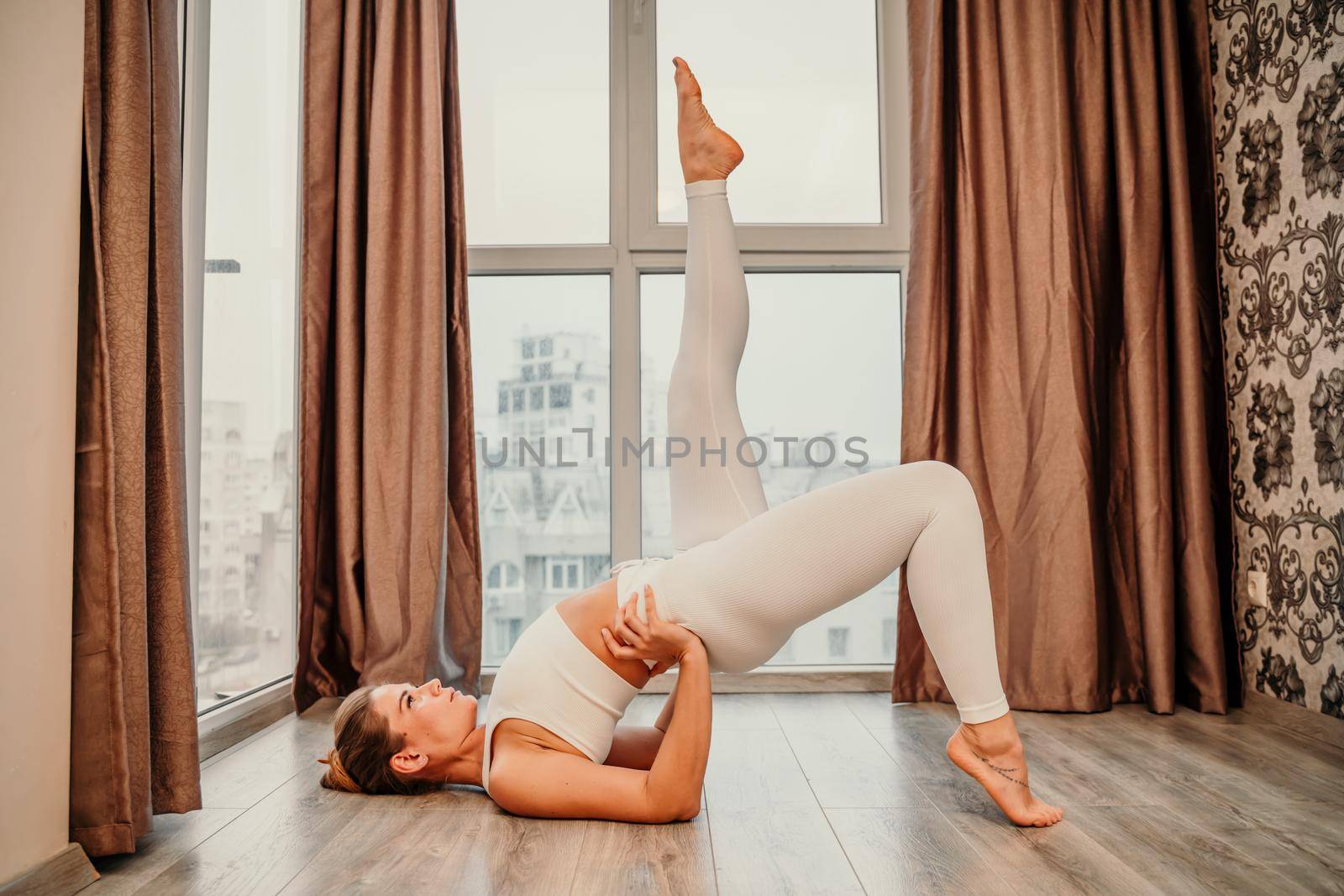 Young athletic attractive woman practicing yoga. Works out at home or in a yoga studio, sportswear, white pants and a full-length top indoors. Healthy lifestyle concept by Matiunina