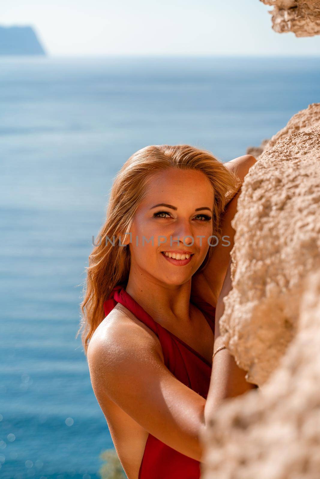 Smiling young woman in a red dress looks at the camera. A beautiful tanned girl enjoys her summer holidays at the sea. Portrait of a stylish carefree woman laughing at the ocean. by Matiunina