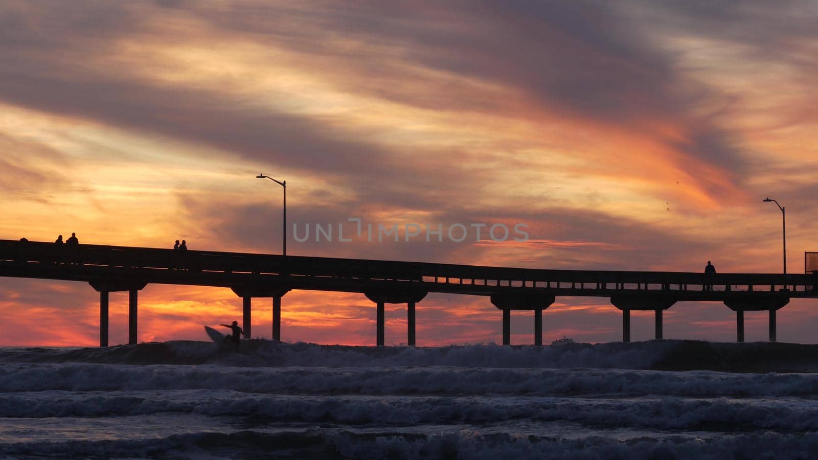 Silhouette of people walking, surfers surfing by pier in sea water. Ocean waves, dramatic sky at sunset. California coast, beach or shore vibes at sundown. Summer seascape seamless looped cinemagraph.
