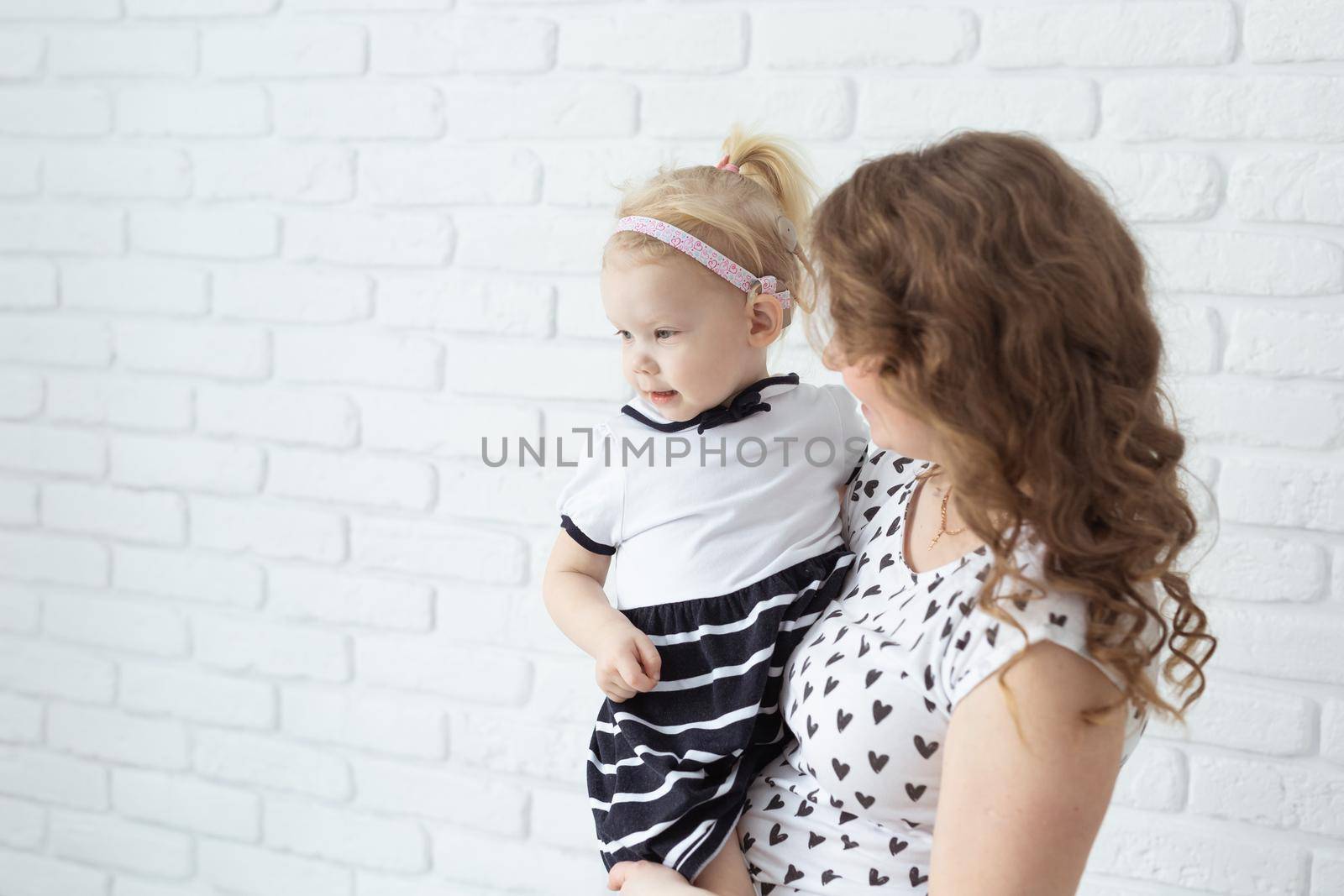 Mother holds her child with hearing aids and cochlear implants on white brick wall with copy space and place for advertising . Deaf and health concept by Satura86