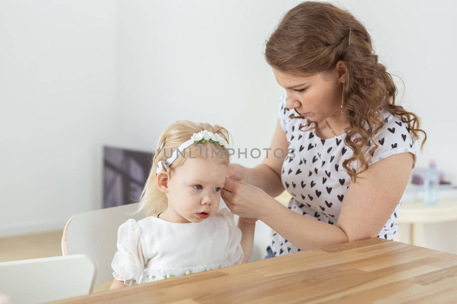 Mother helps her deaf baby daughter putting hearing aid in little girl's ear indoors - cochlear implant and innovative medical technologies in the treatment of deafness by Satura86