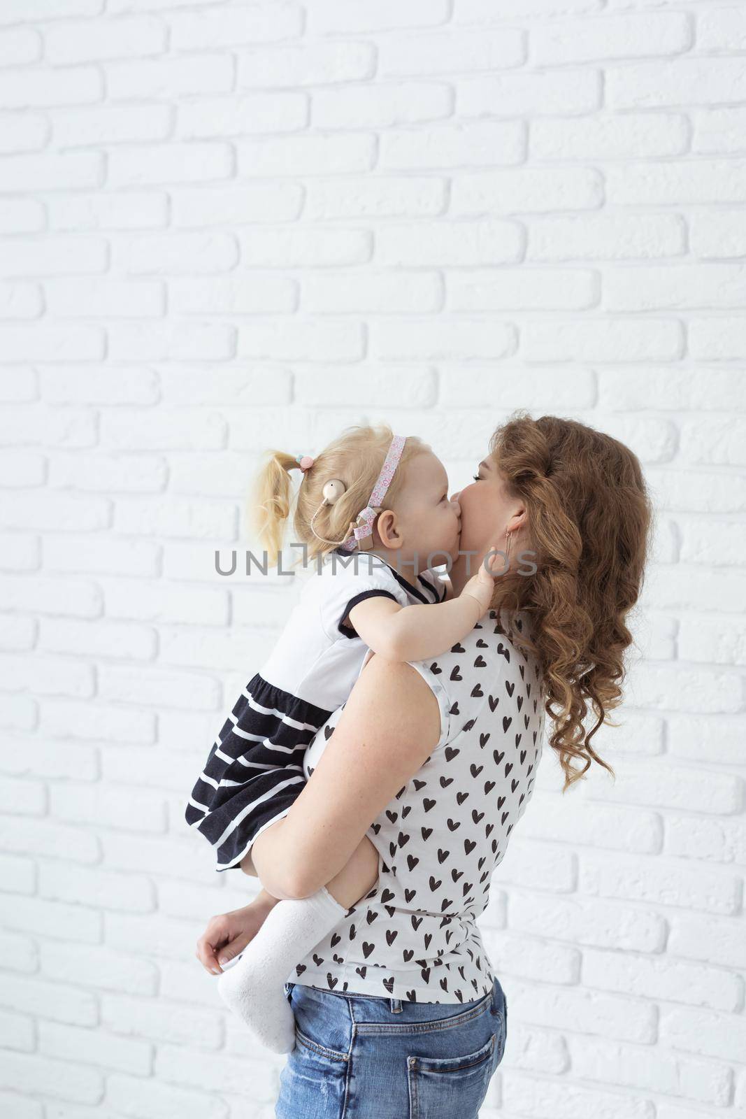 Mother holds and having fun her child with hearing aids and cochlear implants on white brick wall with copy space and place for advertising . Deaf and innovating medicine technologies concept by Satura86