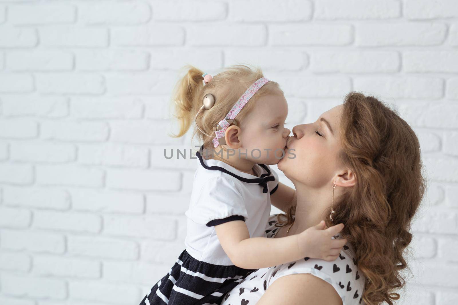 Mother holds and having fun her child with hearing aids and cochlear implants on white brick wall with copy space and place for advertising . Deaf and innovating medicine technologies concept by Satura86