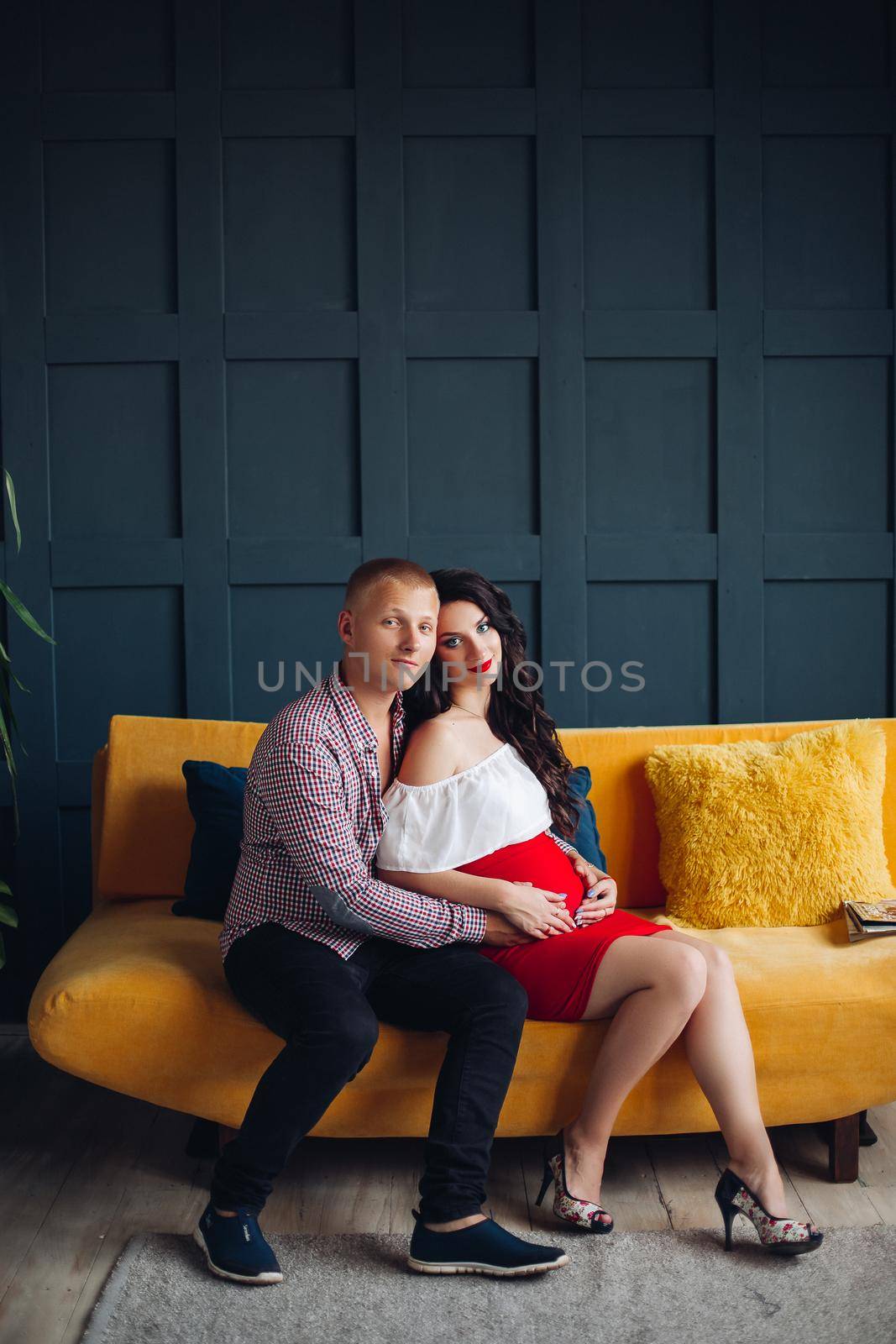Family waiting for baby and loving each other. Elegant and stylish couple sitting on yellow couch and looking at camera. Blonde husband hugging pregnant brunette wife and holding hands on her belly.