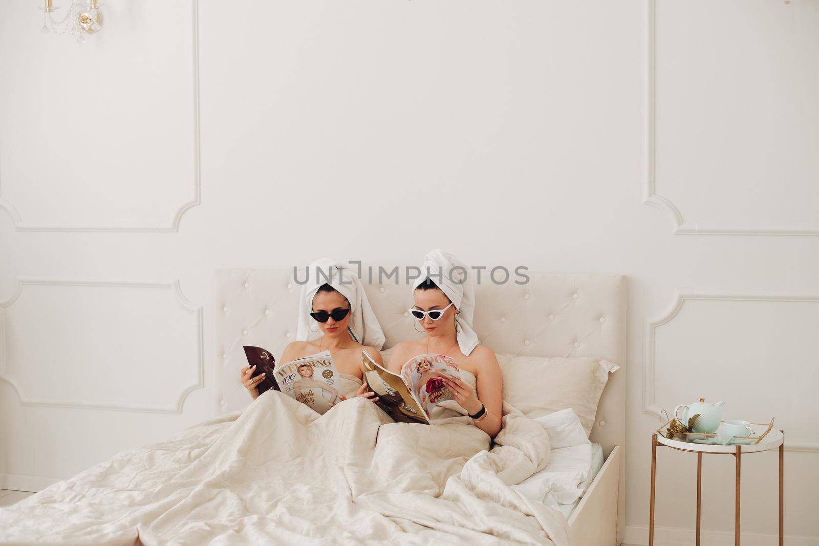 Stock photo of incognito women in bath towels on head and sunglasses holding fashion magazines sitting in bed. Fashion victims in the morning. Fashion divas preparing for the day reading fashion journals in bed.