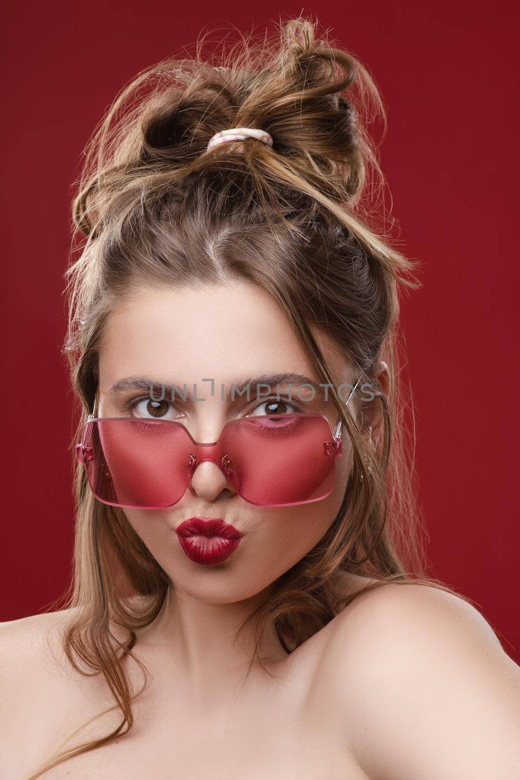 Fashion portrait of stunning young girl with messy hairstyle wearing trendy red or pink sunglasses and red lips. Red color concept. Sunglasses match lips and background.