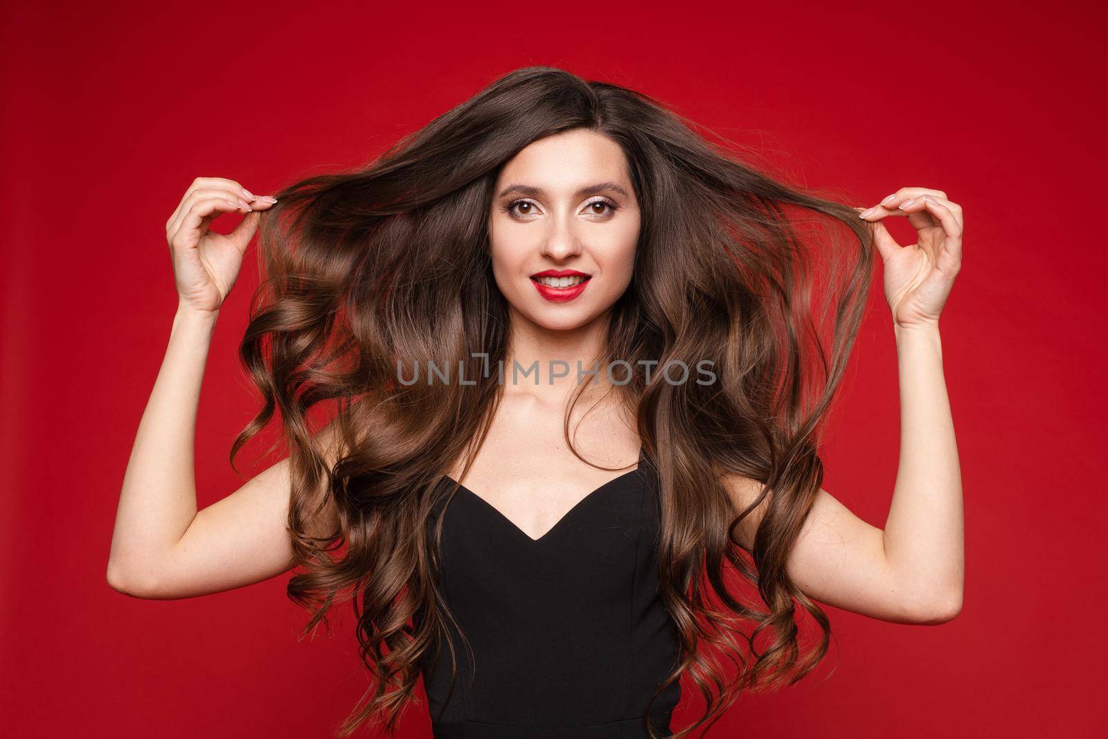 Studio portrait of stunning young caucasian woman with gorgeous long brunette hair in black with red lips. Isolate on red. She is showing beautiful, smooth and healthy hair.