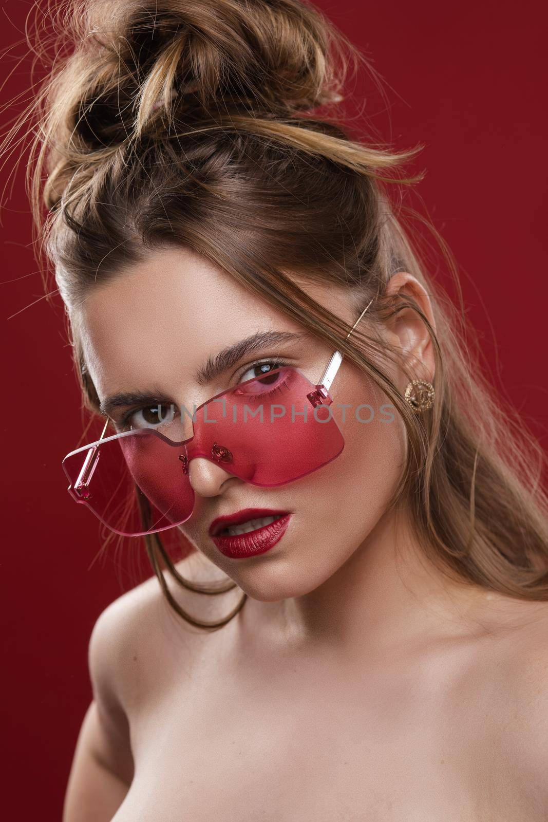 Fashion portrait of stunning young girl with messy hairstyle wearing trendy red or pink sunglasses and red lips. Red color concept. Sunglasses match lips and background.