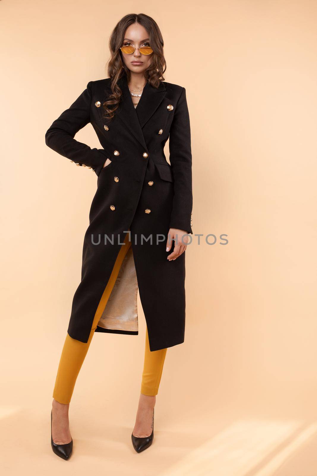 Full length fashionable portrait of gorgeous young brunette woman in trendy sunglasses, black double-breasted coat and black high heels. Wavy hair, looking at camera. Isolate on plain background.