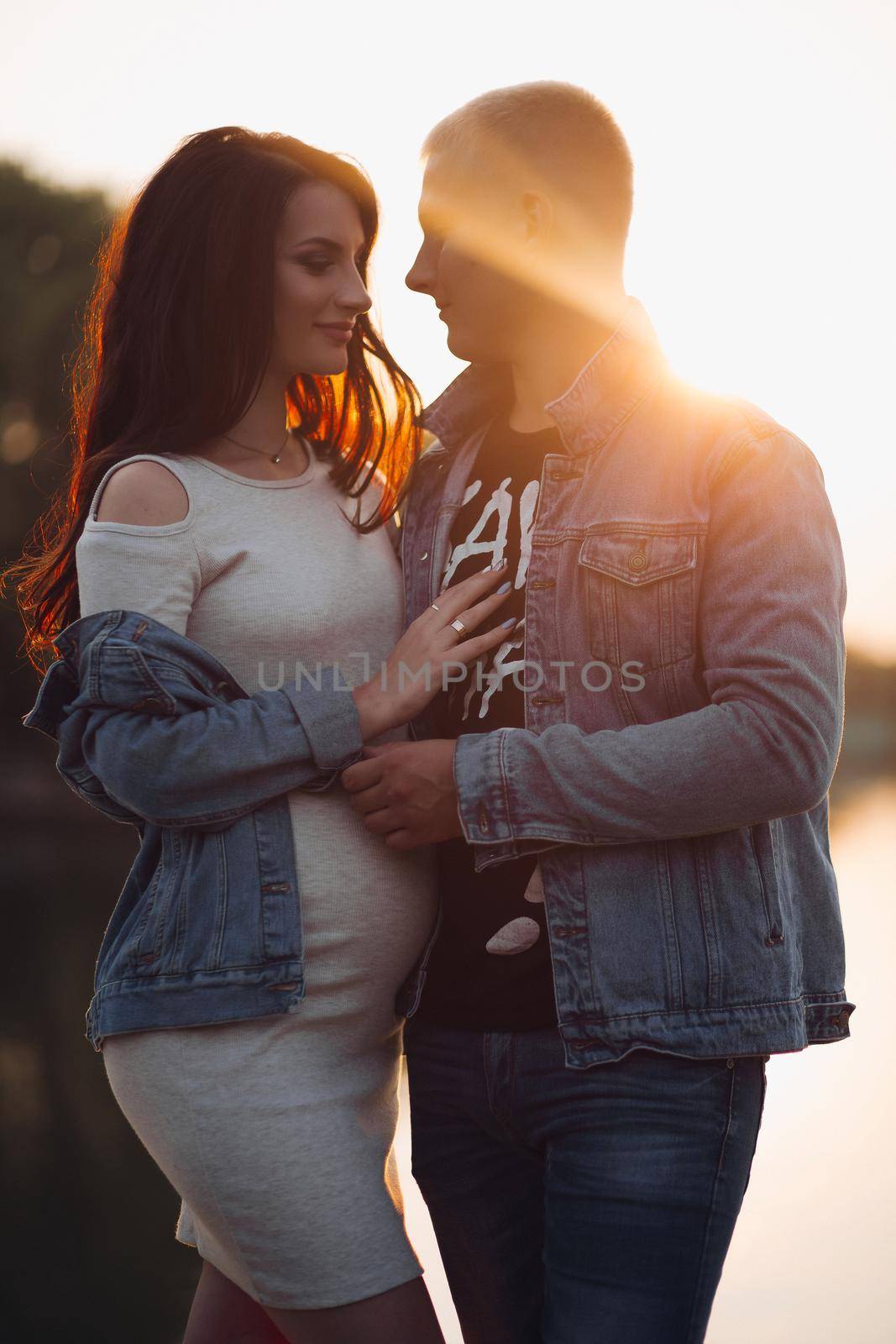 Side view of happy family enjoying moment in rays of sun. Brunette pregnant wife smiling and hugging her blonde husband. Couple of young people in casual clothes relaxing together at end of day.