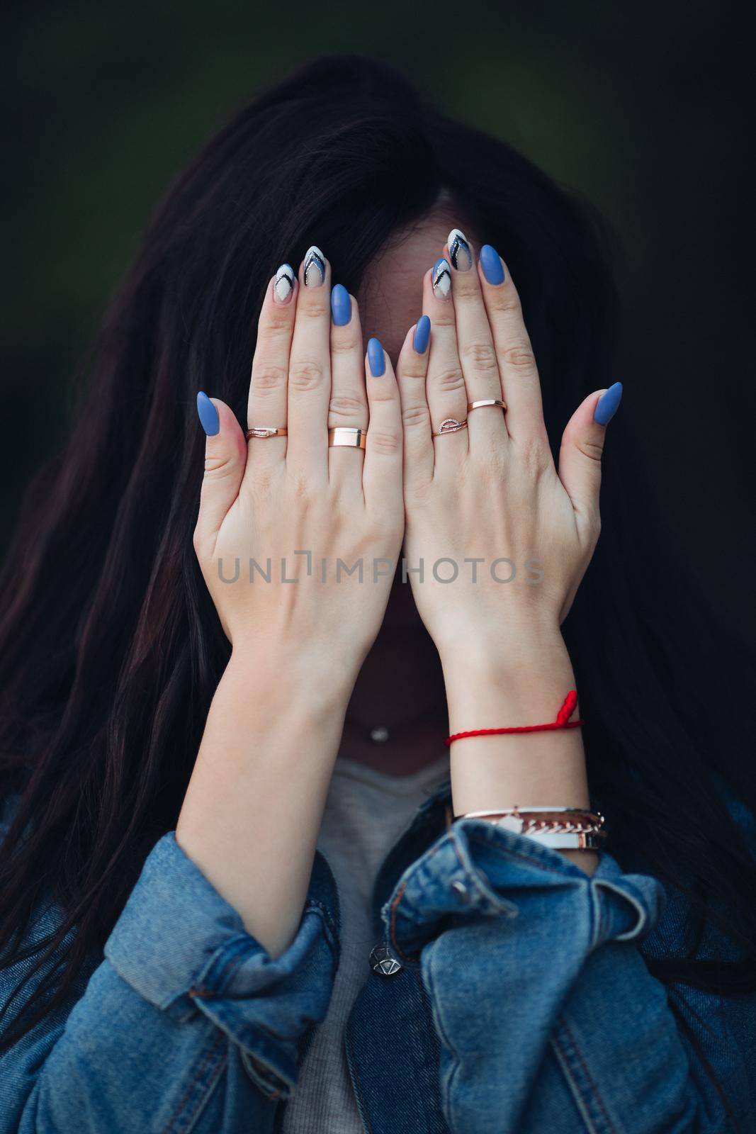 Crop of female hands with elegant blue and white manicure before head. Girl with gorgeous dark long hair hiding face by arms. Woman in jeans jacket holding palms together and wearing rings on fingers.