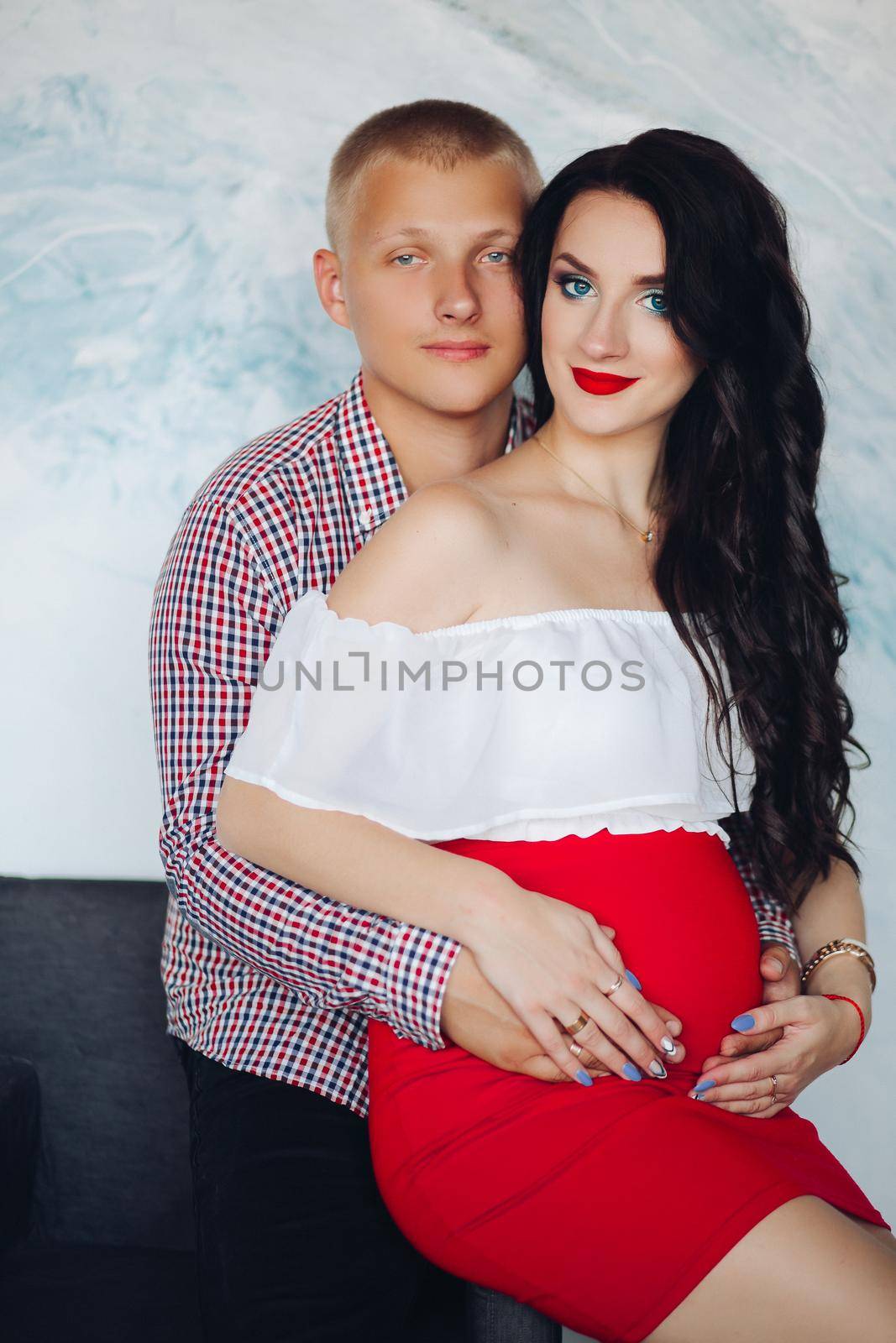 Studio portrait of gorgeous brunette woman with her husband embracing her belly from behind. They looking at camera.