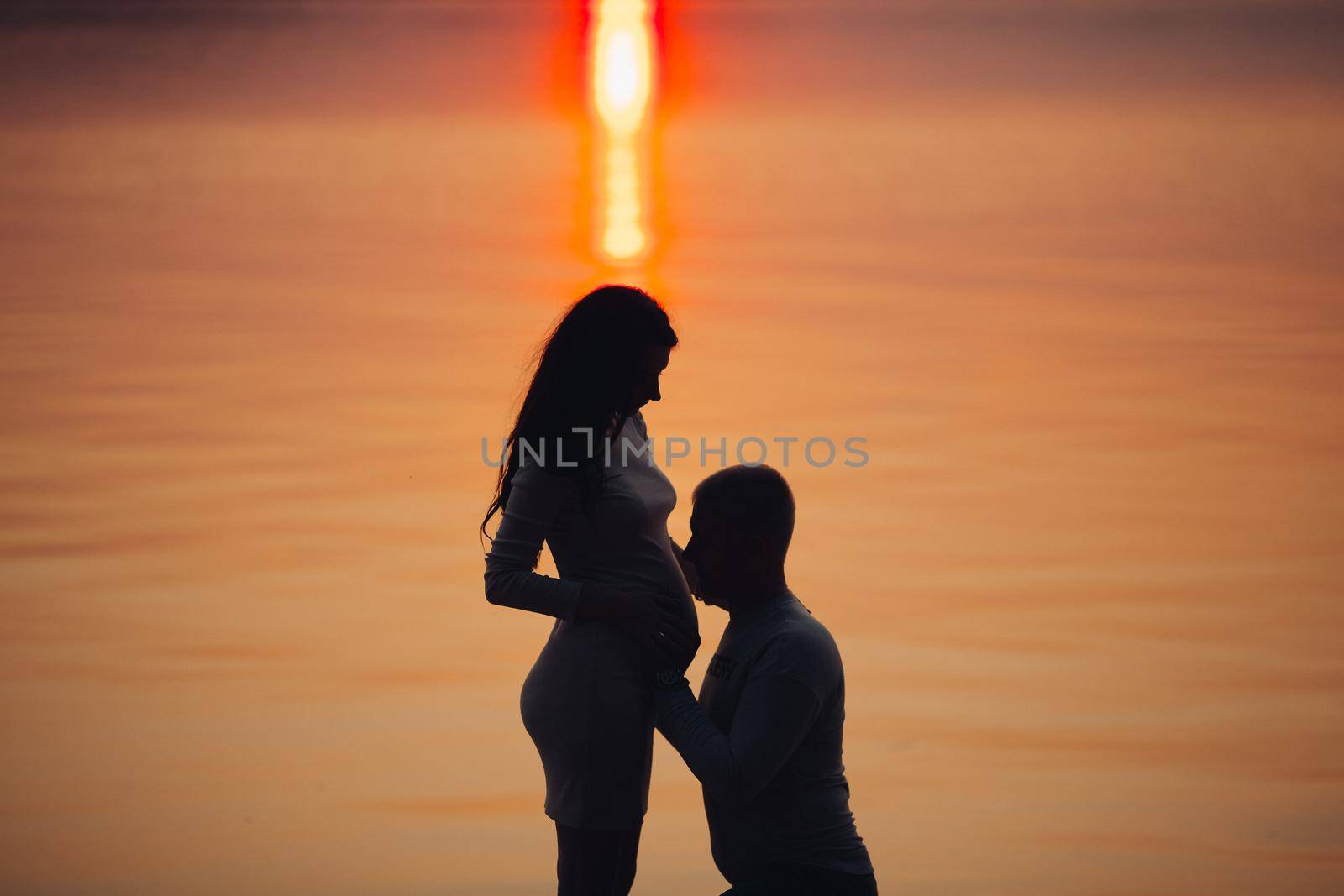 Silhouette of pregnant woman and her husband against sunset by the sea. Man is embracing and kissing her belly. Expecting a baby concept. Evening by the water.