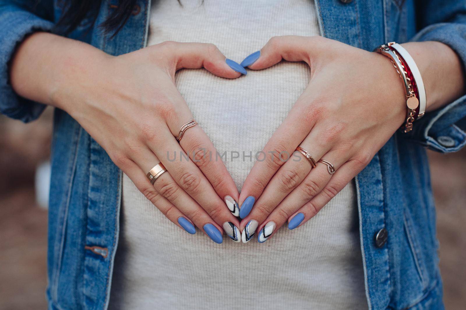 Close up of female belly during pregnancy. Stylish woman in white sweater and jeans jacket holding palms on tummy. Girl wearing bracelets and ring on fingers with manicure gesturing heart by hands.