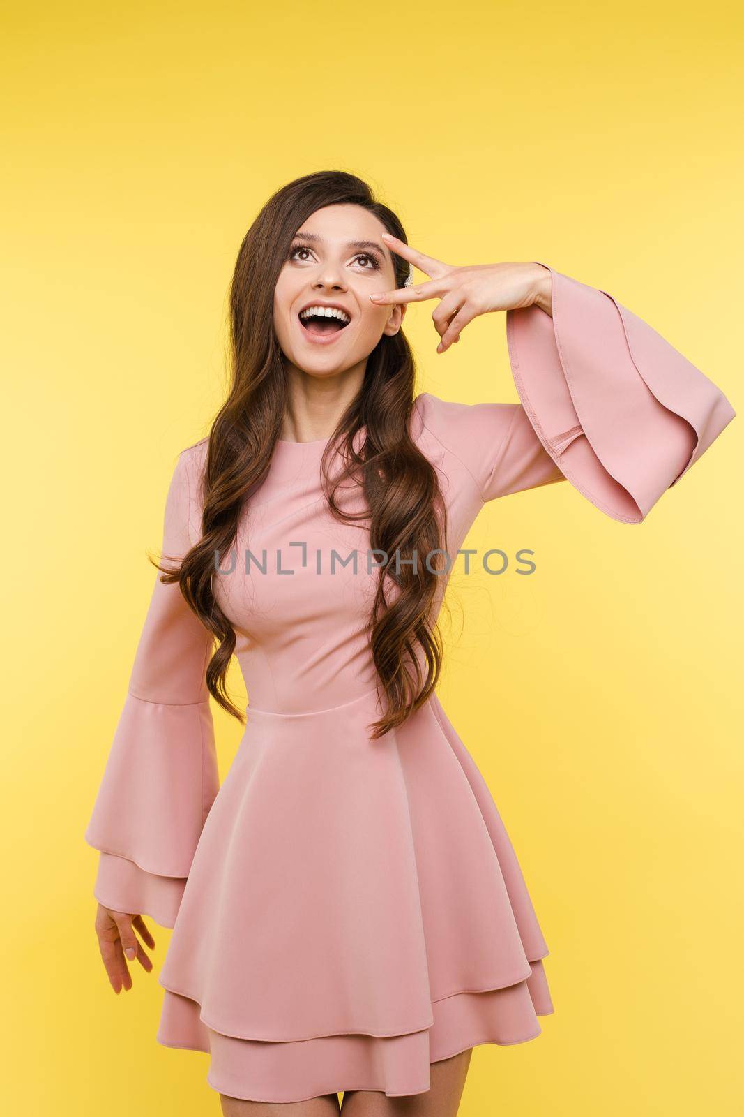Excited stunning lady with long wavy hairstyle wearing trendy pink blouse showing ok gesture looking up with open mouth. Isolate on yellow.