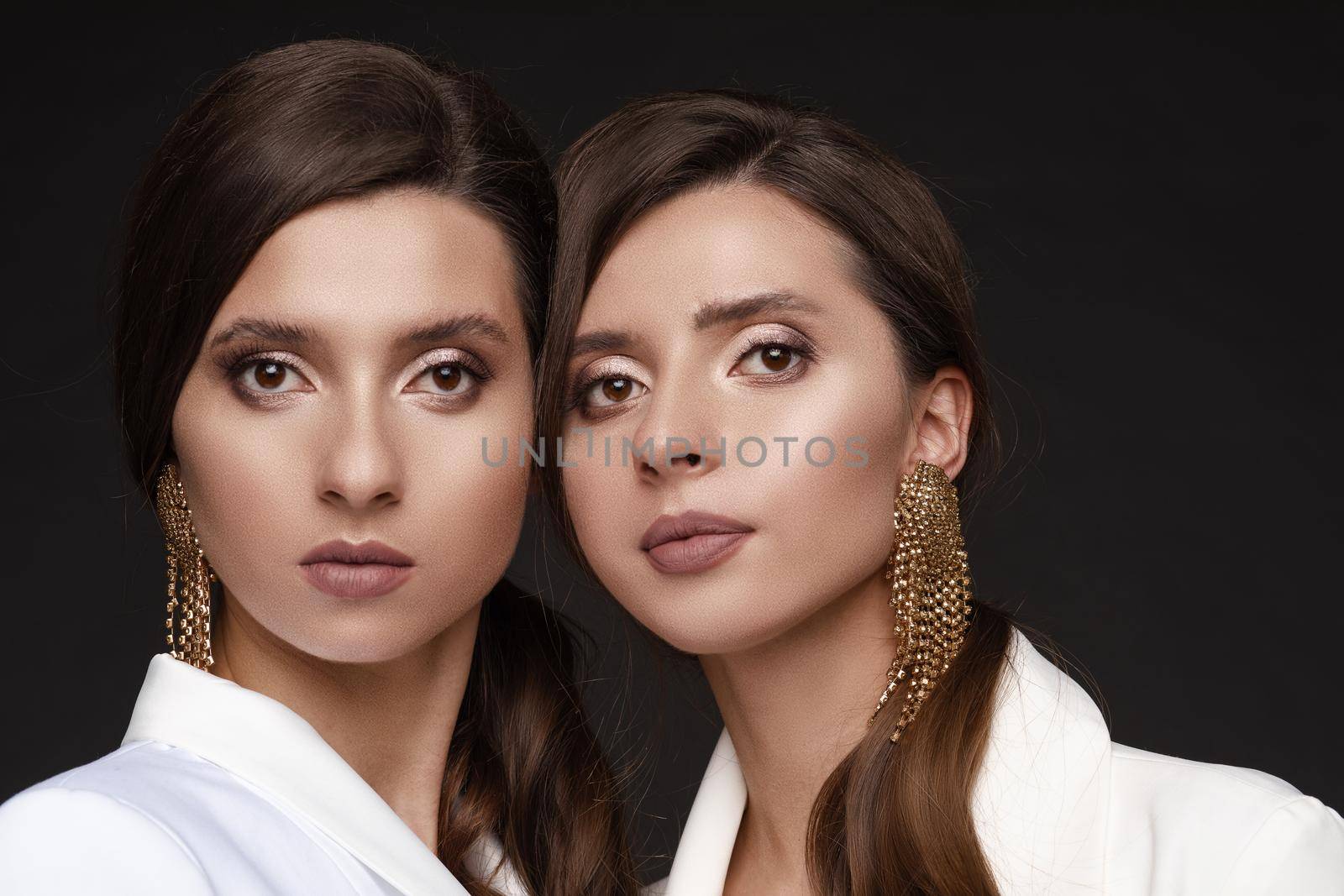 Portrait of two beautiful sisters with elegant make up. Brunette twins with long hair standing close to each other and looking seriously. Young models with big earrings posing together at camera.