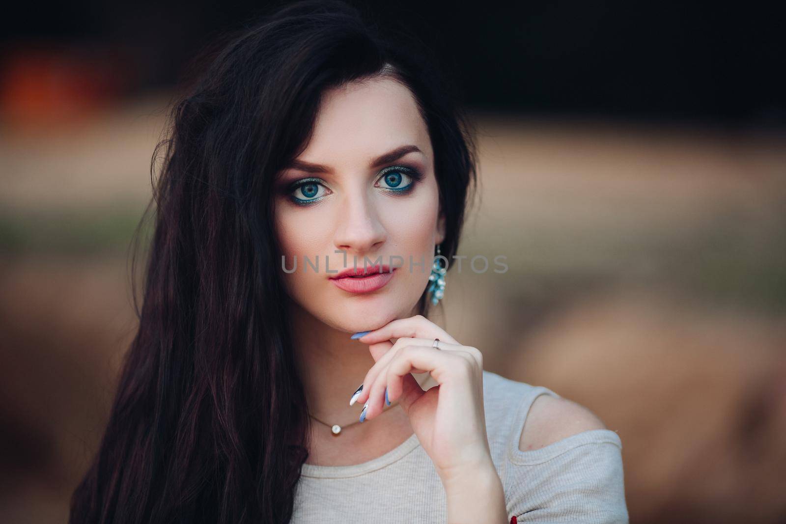 Portrait of attractive young woman with make up looking at camera. Beautiful gorgeous girl with long dark hair holding her hand near face. Crop of elegant pretty lady with blue eyes and full lips.