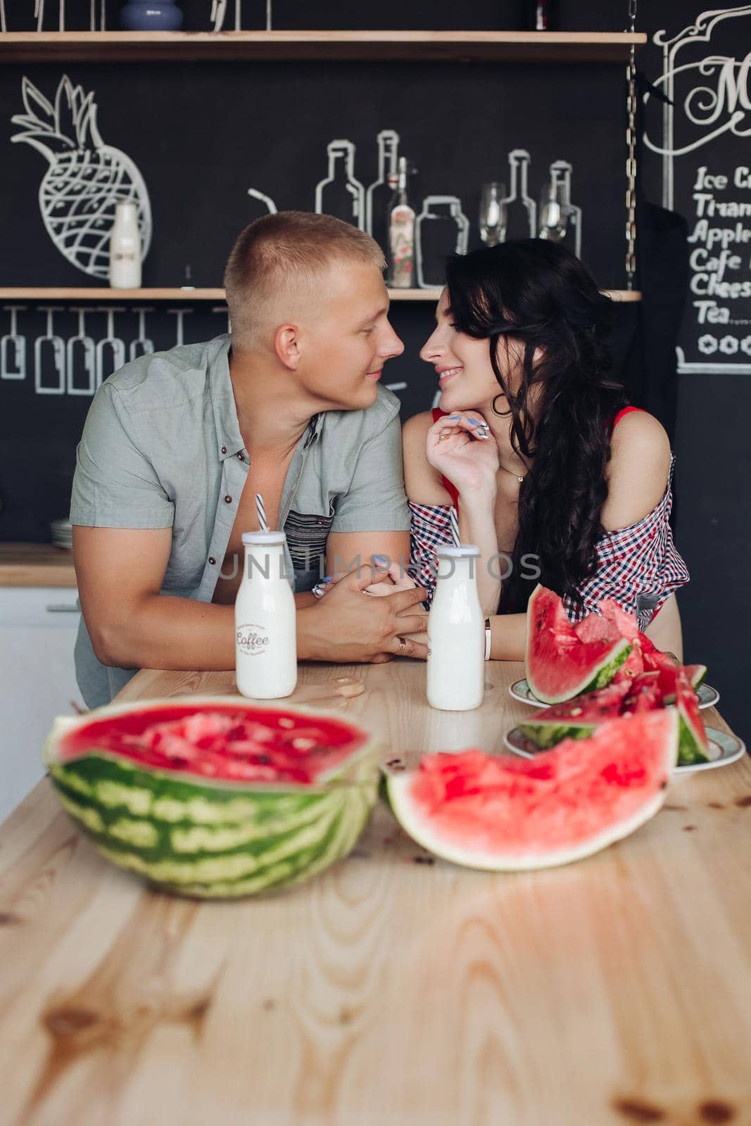 Front view of beautiful pair standing at kitchen, looking at each other and smiling. Lovely wife and husband eating watermelon and drinking yogurt at home. Concept of love and happiness.