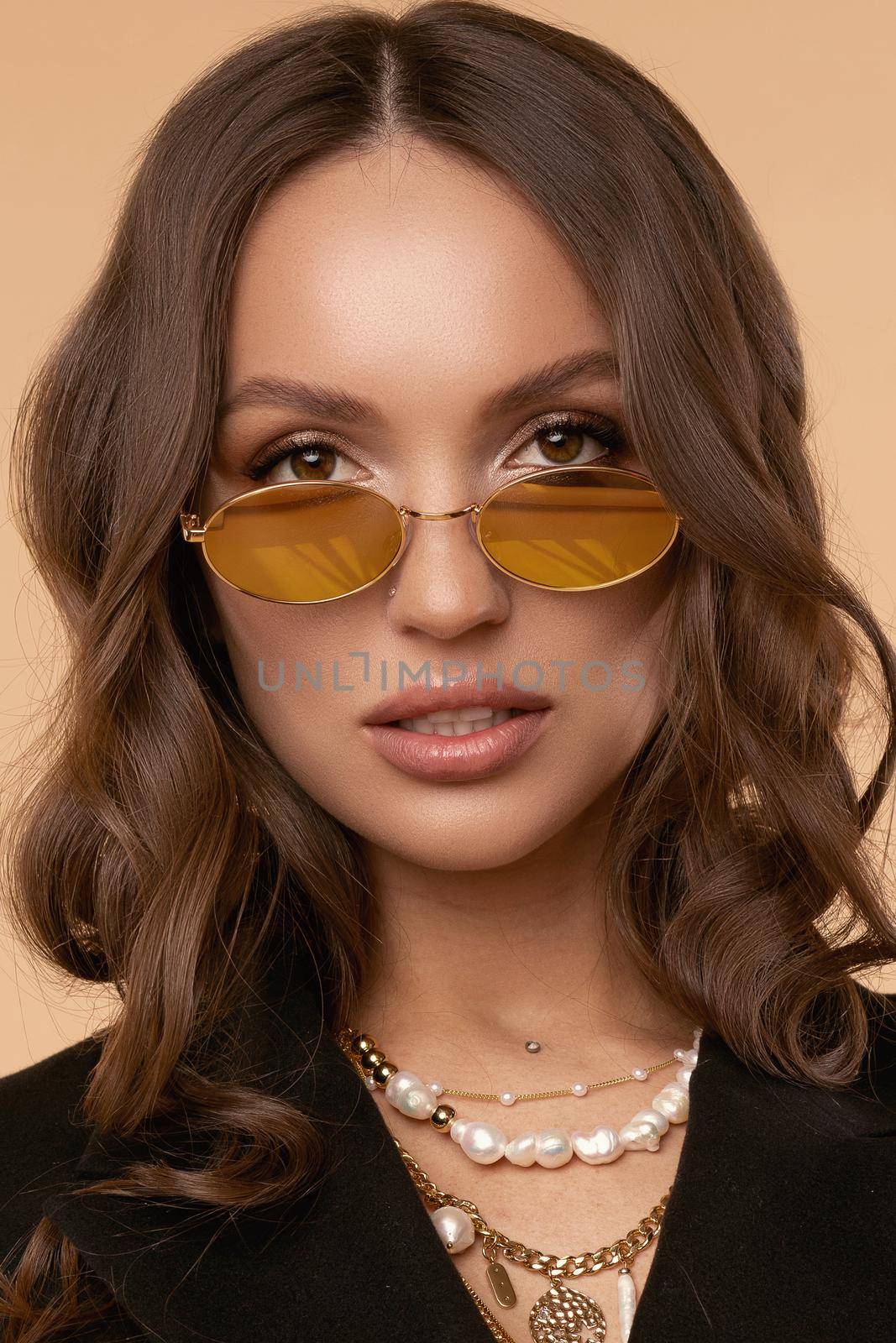 Brunette model with curly hair standing with slightly open mouth. Surprised young girl in sunglasses and black jacket posing on light studio background. Gorgeous lady wearing jewelries on neck.