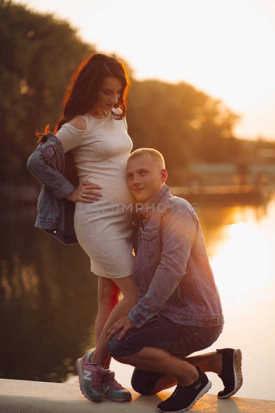 Full length of delighted man in denim jacket listening and embracing his future baby inside his wife s belly. They are in beautiful park by the river or lake at sunset.