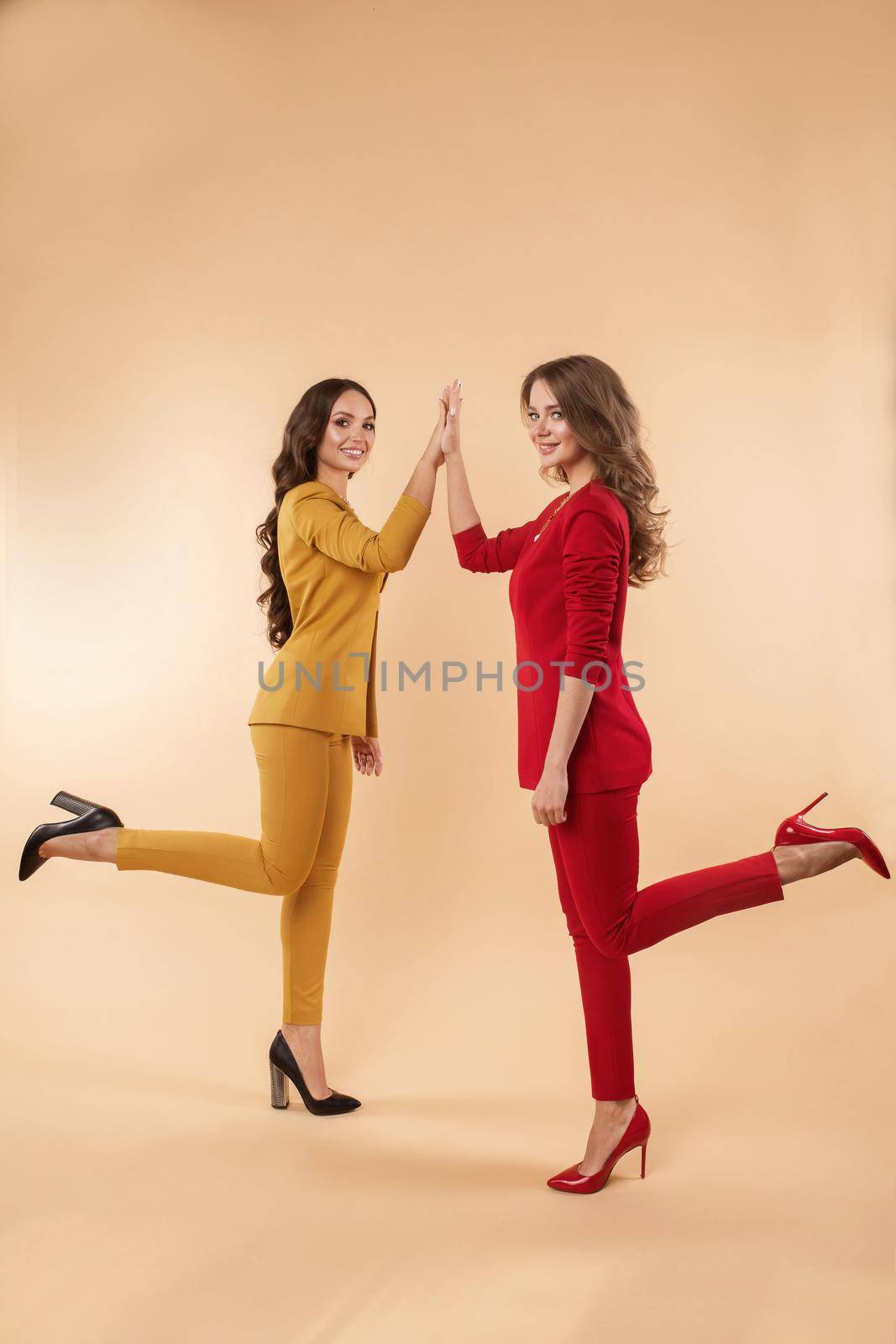 Full length side view of two elegant brunette ladies in stylish bright red and yellow suits and high heels giving high five to each other, smiling at camera. They are cooperating, working together or having an idea. Agreement concept.