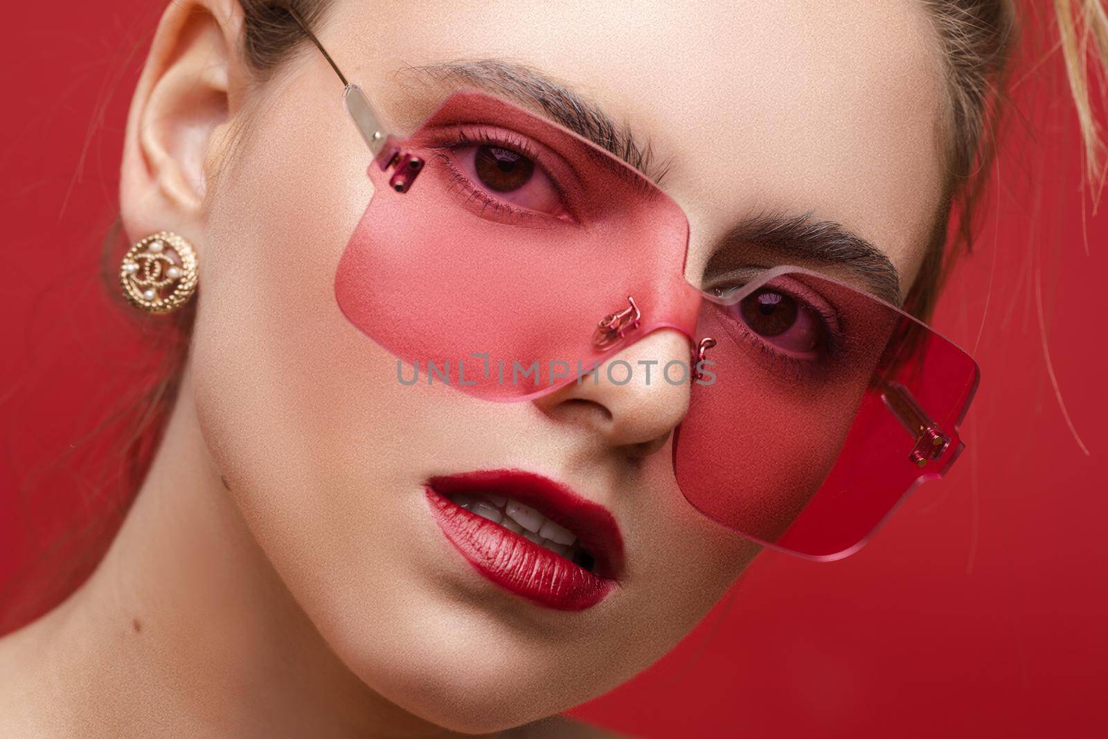 Fashion portrait of stunning young girl with messy hairstyle wearing trendy red or pink sunglasses and red lips. Red color concept. Sunglasses match lips and background.