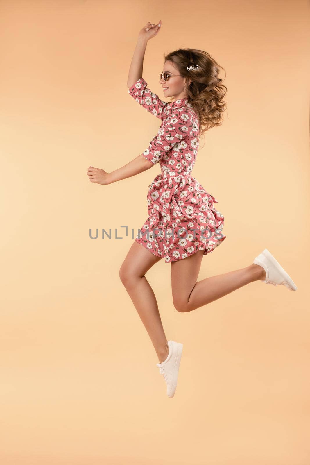 Side view of crazy girl wearing pink floral dress and white sneakers jumping and laughing in studio. Attractive young female in glasses enjoying warm weather and stylish outfit. Concept of happiness.
