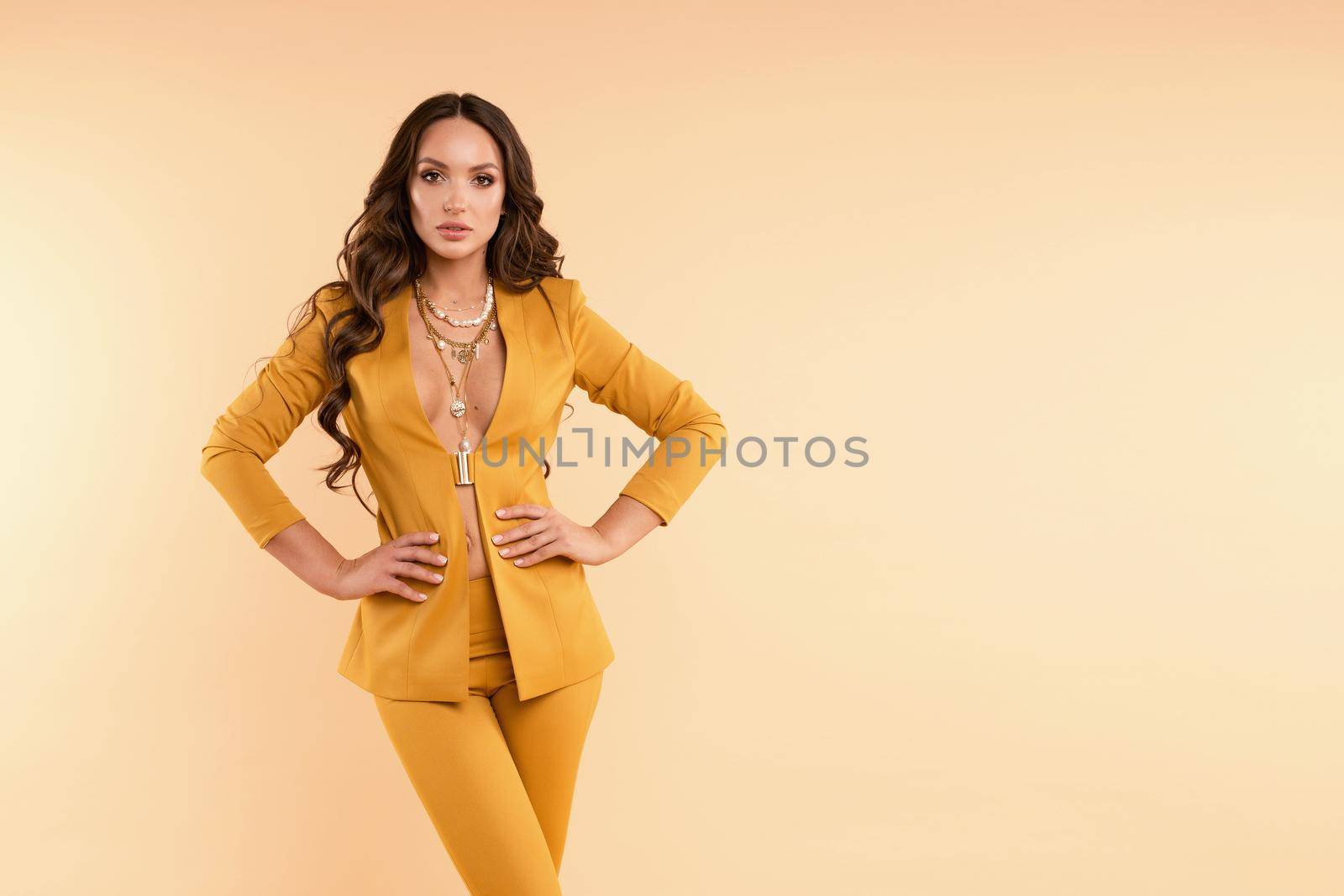 Gorgeous girl with curly hair standing on light studio background. Beautiful brunette lady in yellow elegant costume looking seriously at camera. Professional model holding hand on waist and posing.