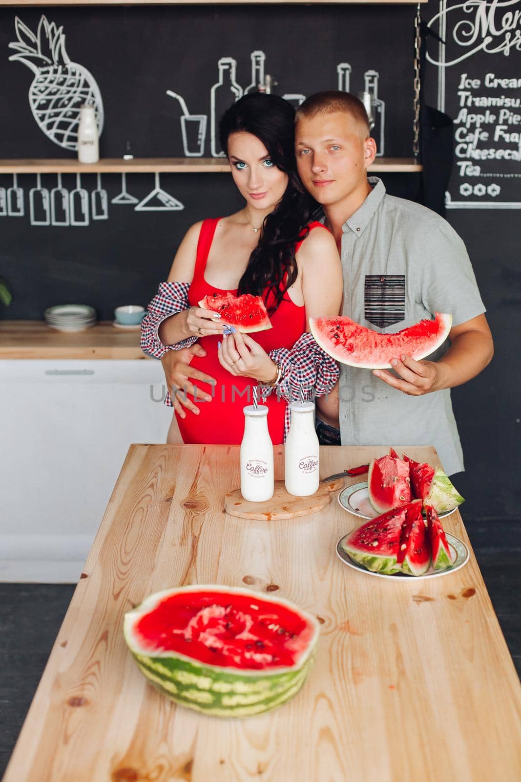 Young husband hugging his beautiful pregnant wife in red top and shirt. They holding sliced watermelon at wooden table in modern kitchen.