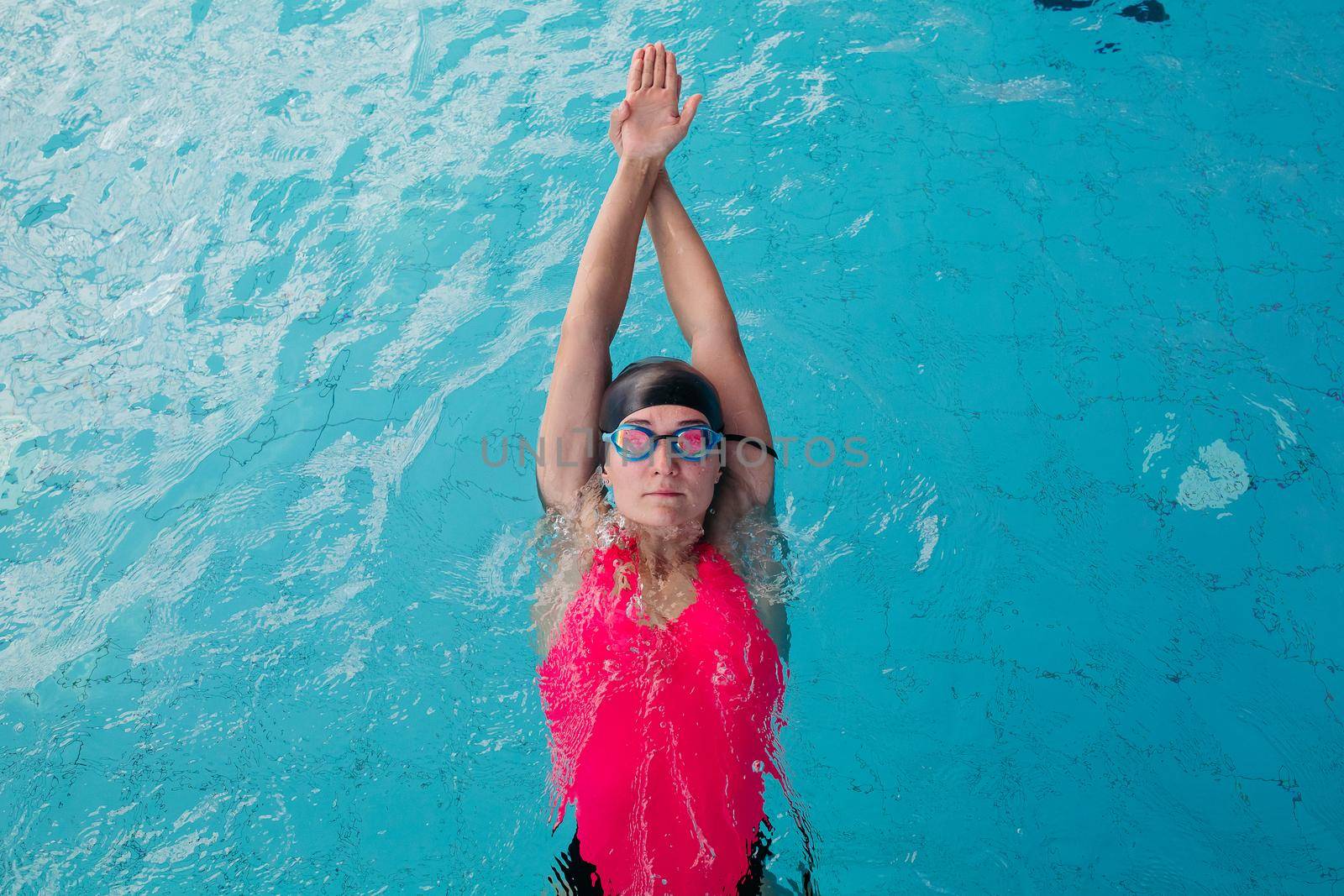 View from above of young sporty girl in goggles and cap swimming in the blue water pool. Woman concentrated swimming forward, wearing in pink swimsuit, engaged in sports. Concept of water sport.