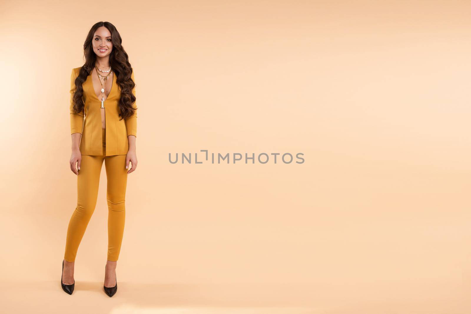 Gorgeous girl with curly hair standing on light studio background. Beautiful brunette lady in yellow elegant costume looking seriously at camera. Professional model holding hand on waist and posing.