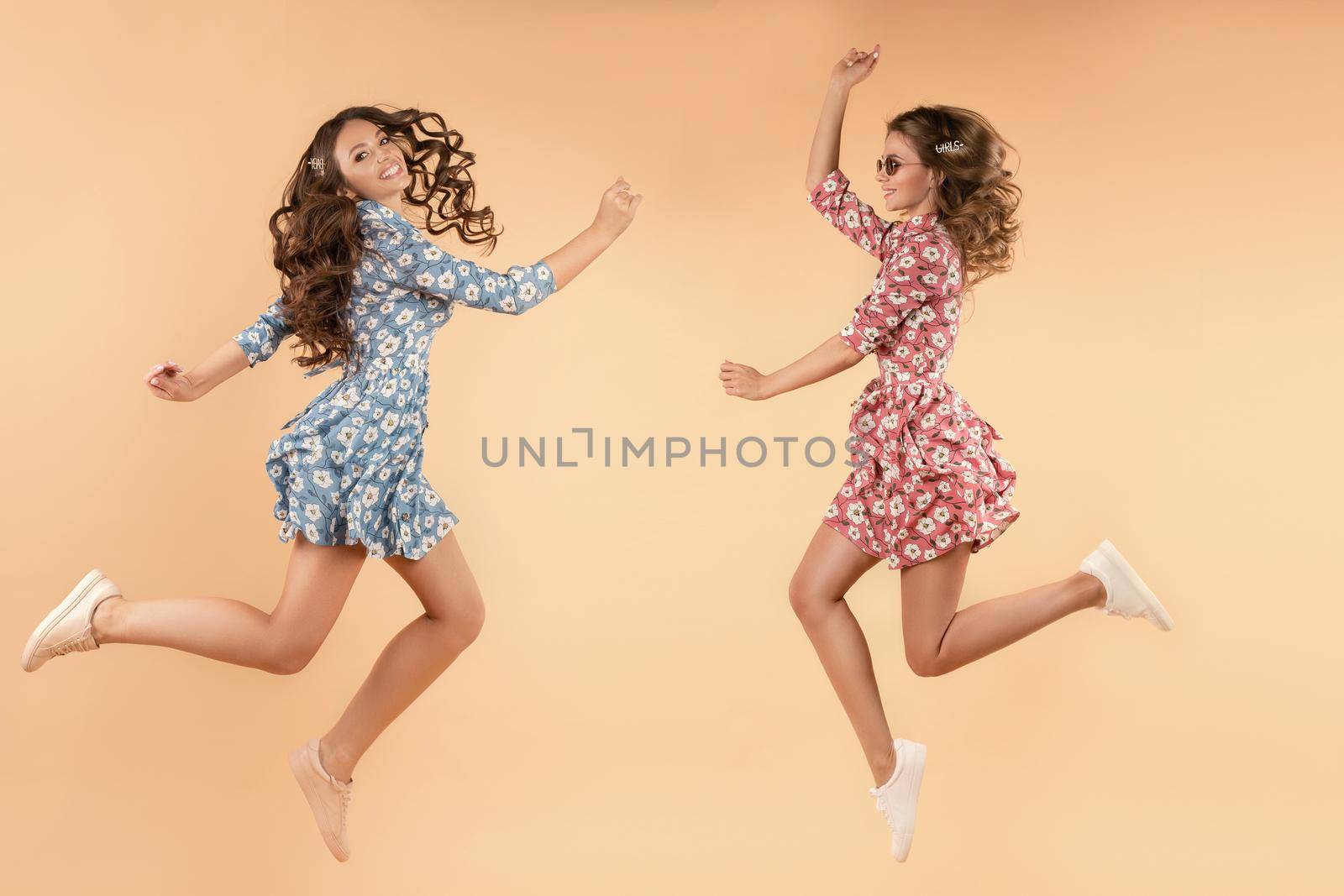 Two beautiful young girls in casual clothes having fun in studio. Professional brunette models with curly hair posing in action. Side view of happy sisters in red and blue dresses jumping and smiling.