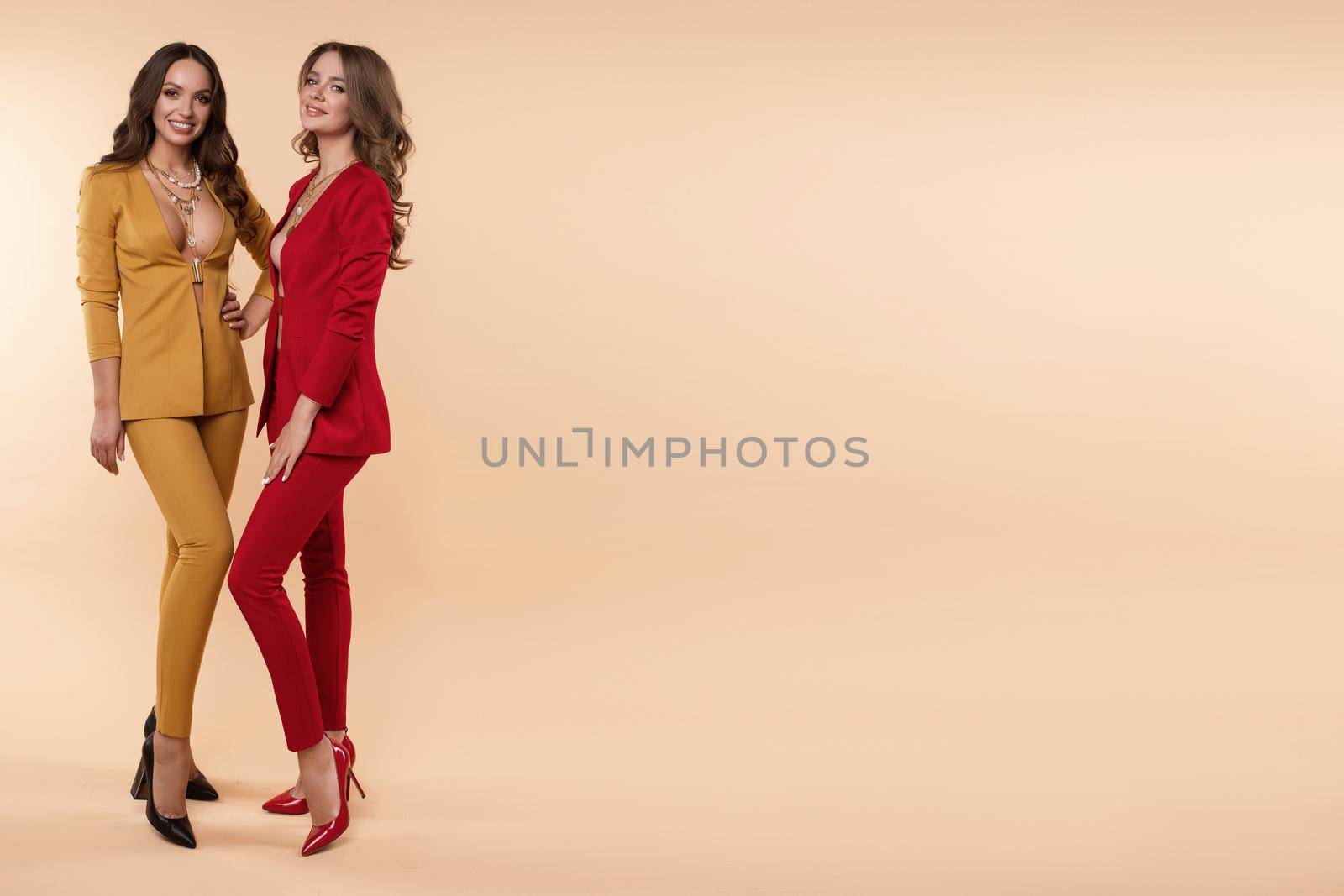 Full length studio portrait of stylish elegant brunette women in red and yellow office suits and high heels. They holding folded arms and looking at camera, posing on flat one-colored background. Studio shot.