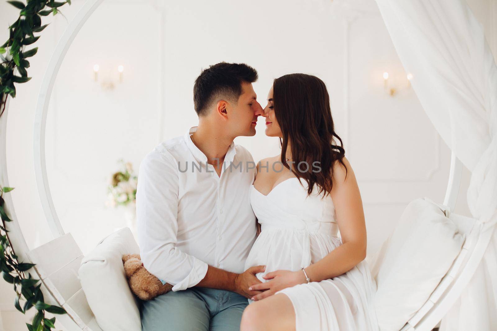 Beautiful couple of young people sitting on white swing with green leaves. Attractive woman and man looking at each other and holding hands together on female belly. Happy family waiting for baby.