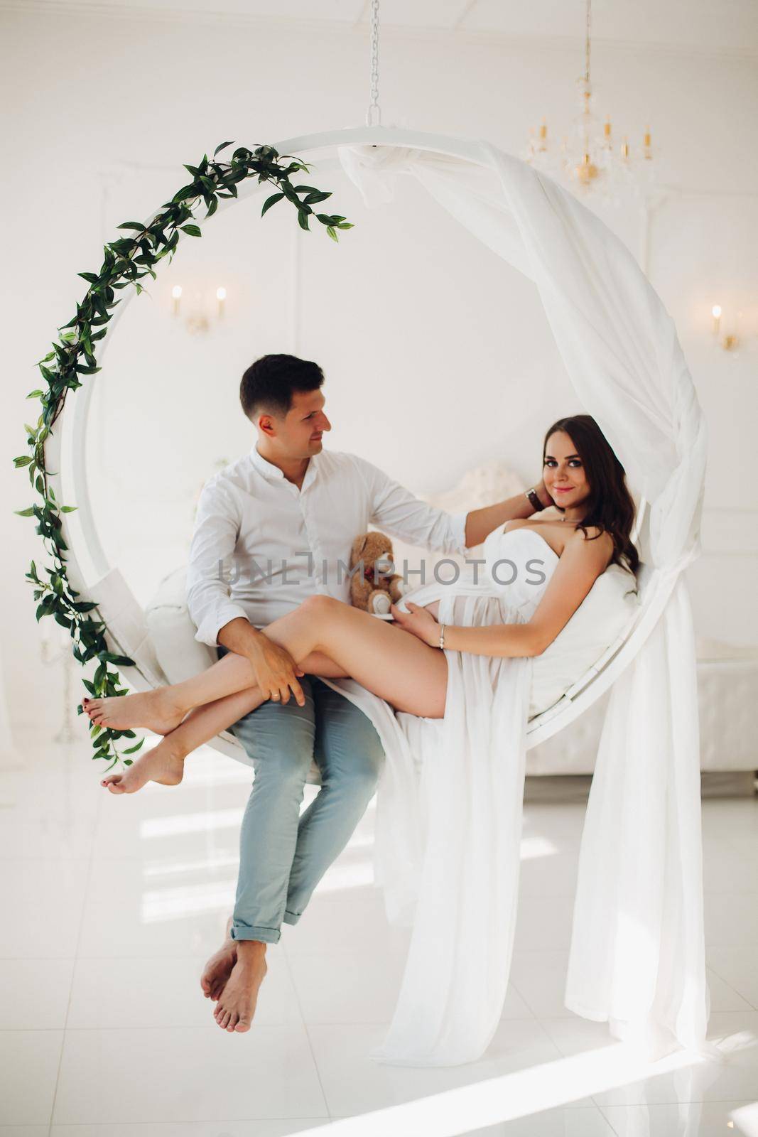 Beautiful couple of young people sitting on white swing with green leaves. Attractive woman and man looking at each other and holding hands together on female belly. Happy family waiting for baby.