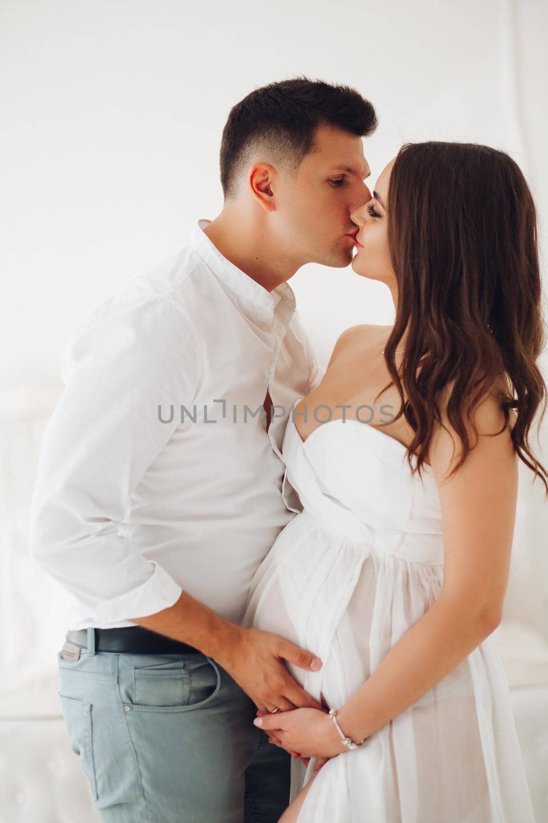 Happy future mother and father hugging each other, looking at camera and smiling. Attractive pregnant woman in white dress awaiting little baby. Concept of pregnancy and happiness.