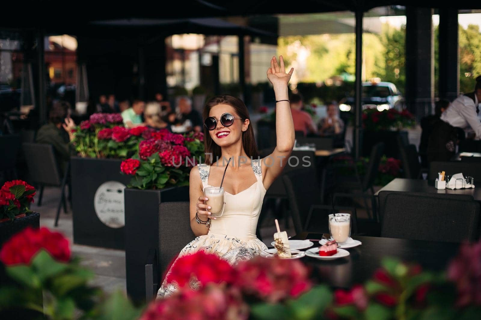 Portrait of a beautiful joyful brunette woman in a cafe. She is dressed in a sumptuous stylish pink dress and stylish glasses. He handsily lifts his hand up and holds a cocktail. Concept of a joyful woman in a cafe.