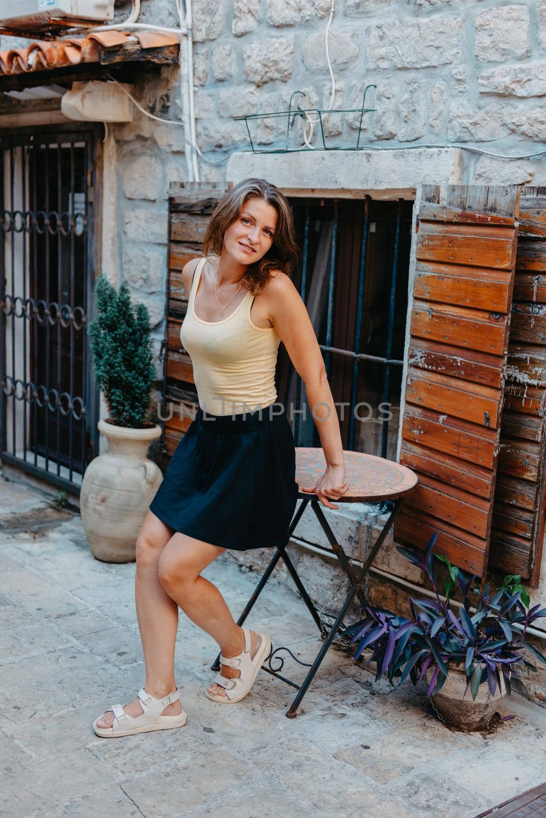 Girl Tourist Walking Through Ancient Narrow Street On A Beautiful Summer Day In MEDITERRANEAN MEDIEVAL CITY, OLD TOWN BUDVA, MONTENEGRO. Young Beautiful Cheerful Woman Walking On Old Street At Tropical Town. Pretty Girl Looking At You And Smiling by Andrii_Ko